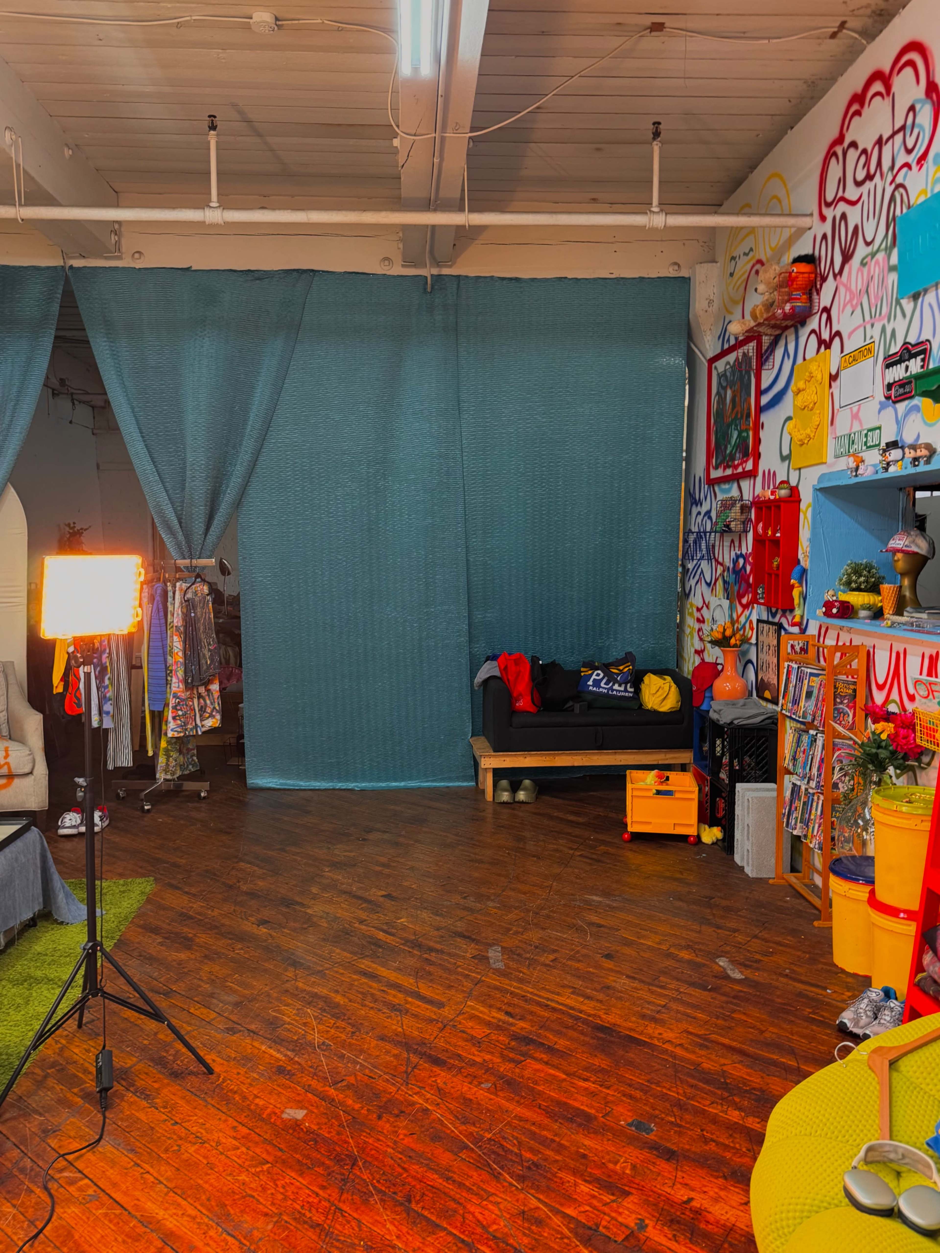 The scene shows a vibrant creative space featuring a couch, colorful wall art, and a studio light, with a teal curtain partially dividing the area.