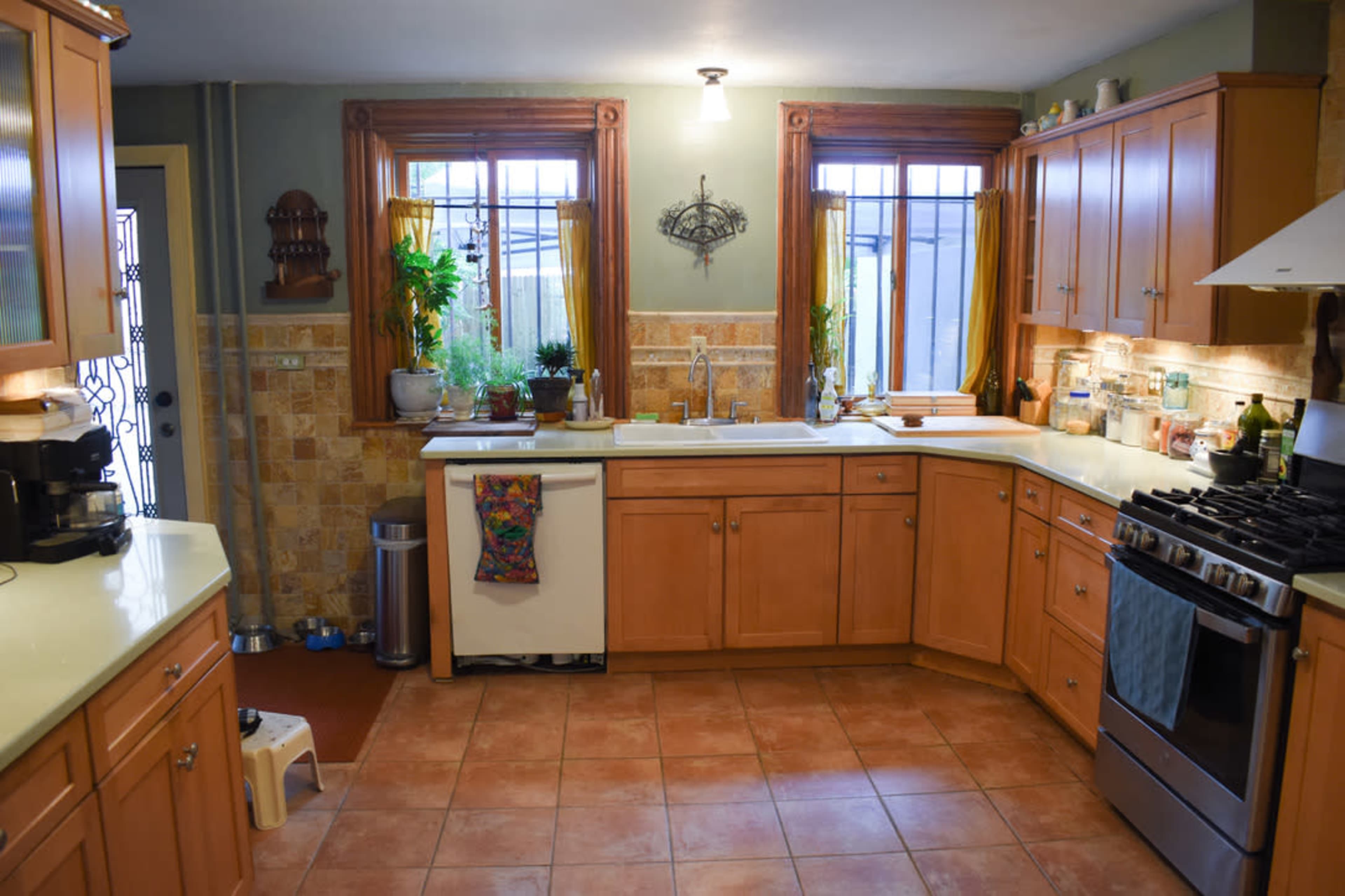 A modern kitchen features wooden cabinets, tiled countertops, and natural light streaming through two windows.