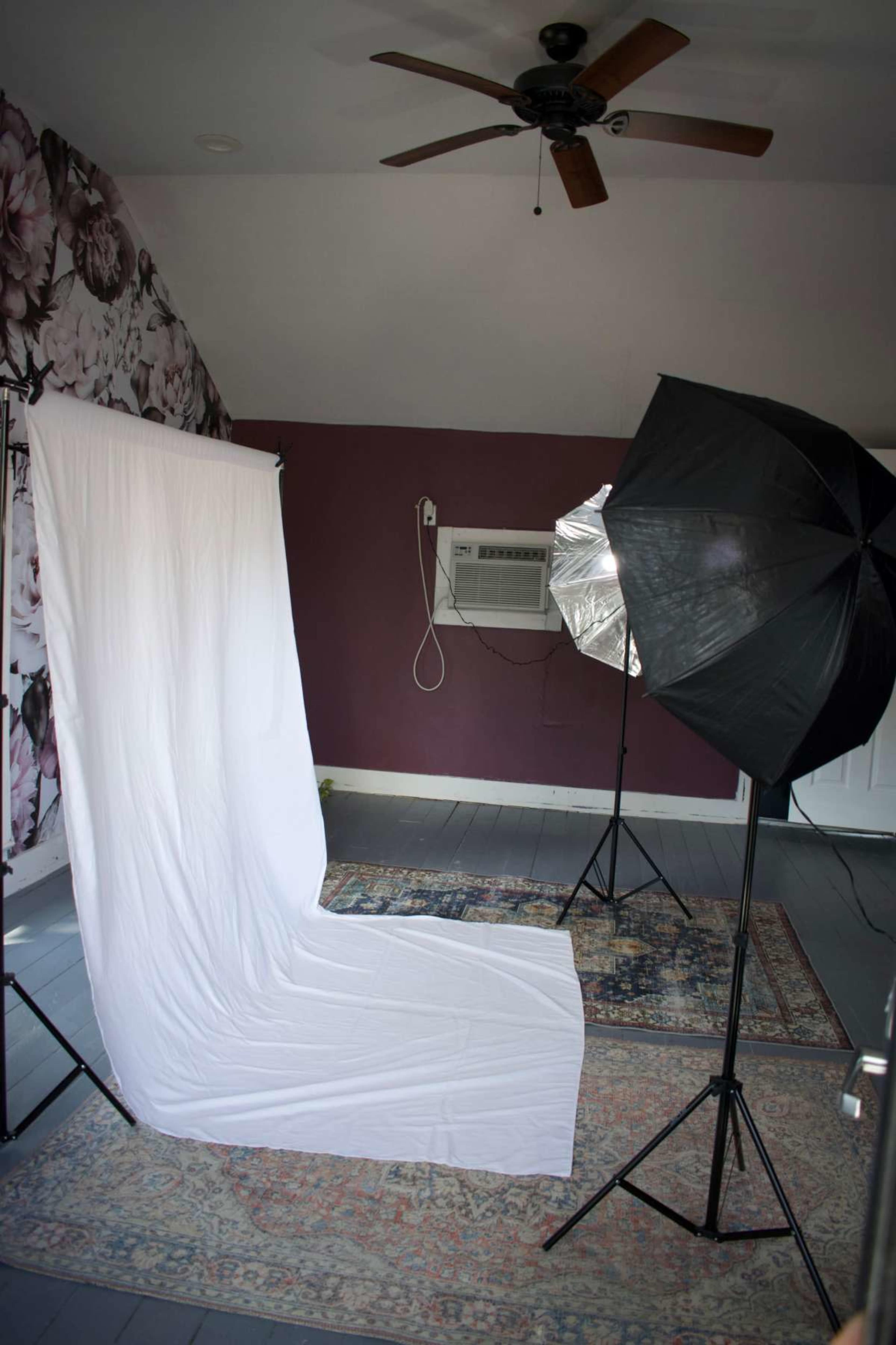 The image shows a photography setup with a white backdrop, a softbox light, and a patterned rug in a room with floral wallpaper and a ceiling fan.