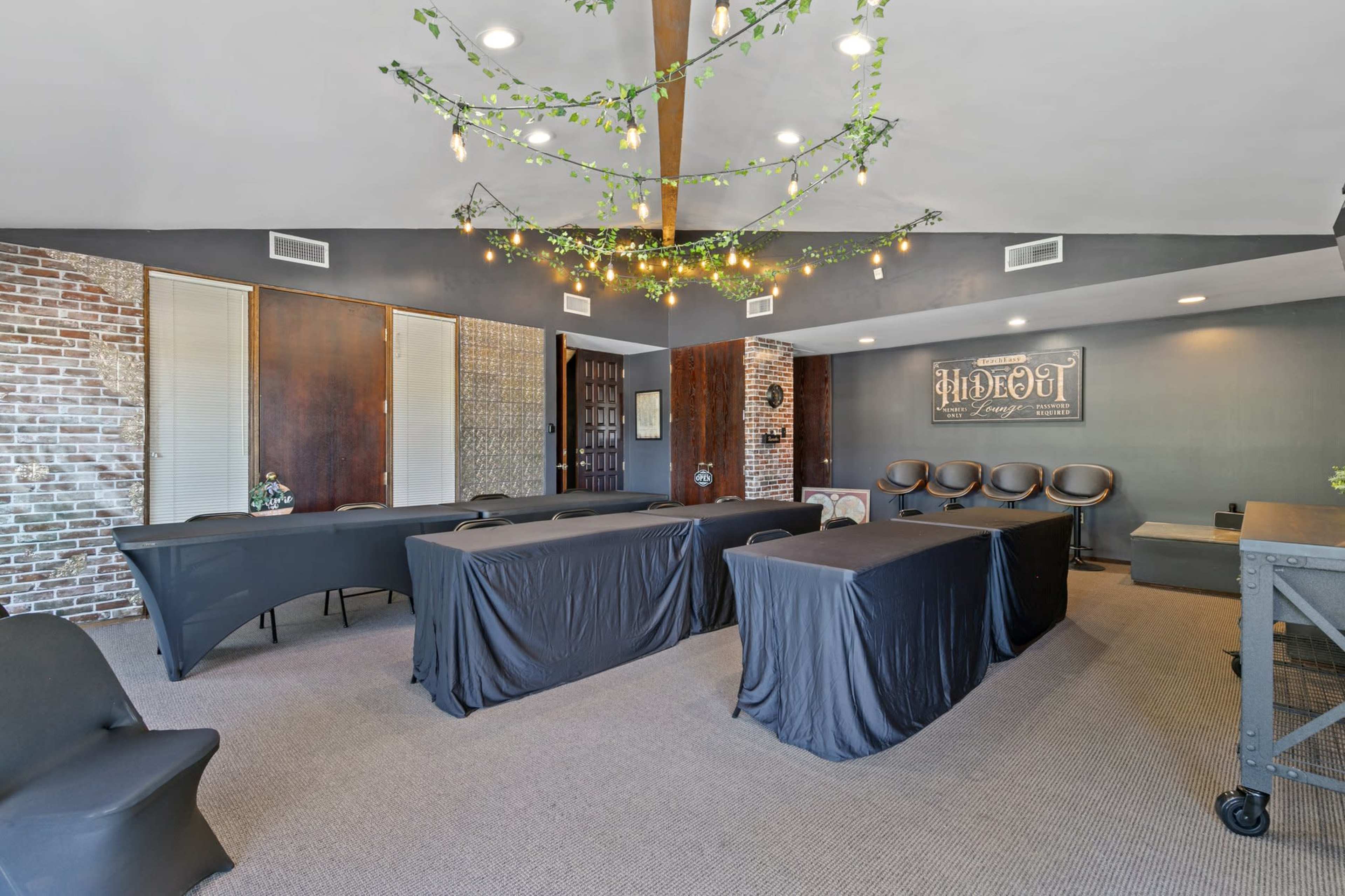 A conference room features several tables covered with black linens, surrounded by chairs, and decorated with hanging greenery and lights.