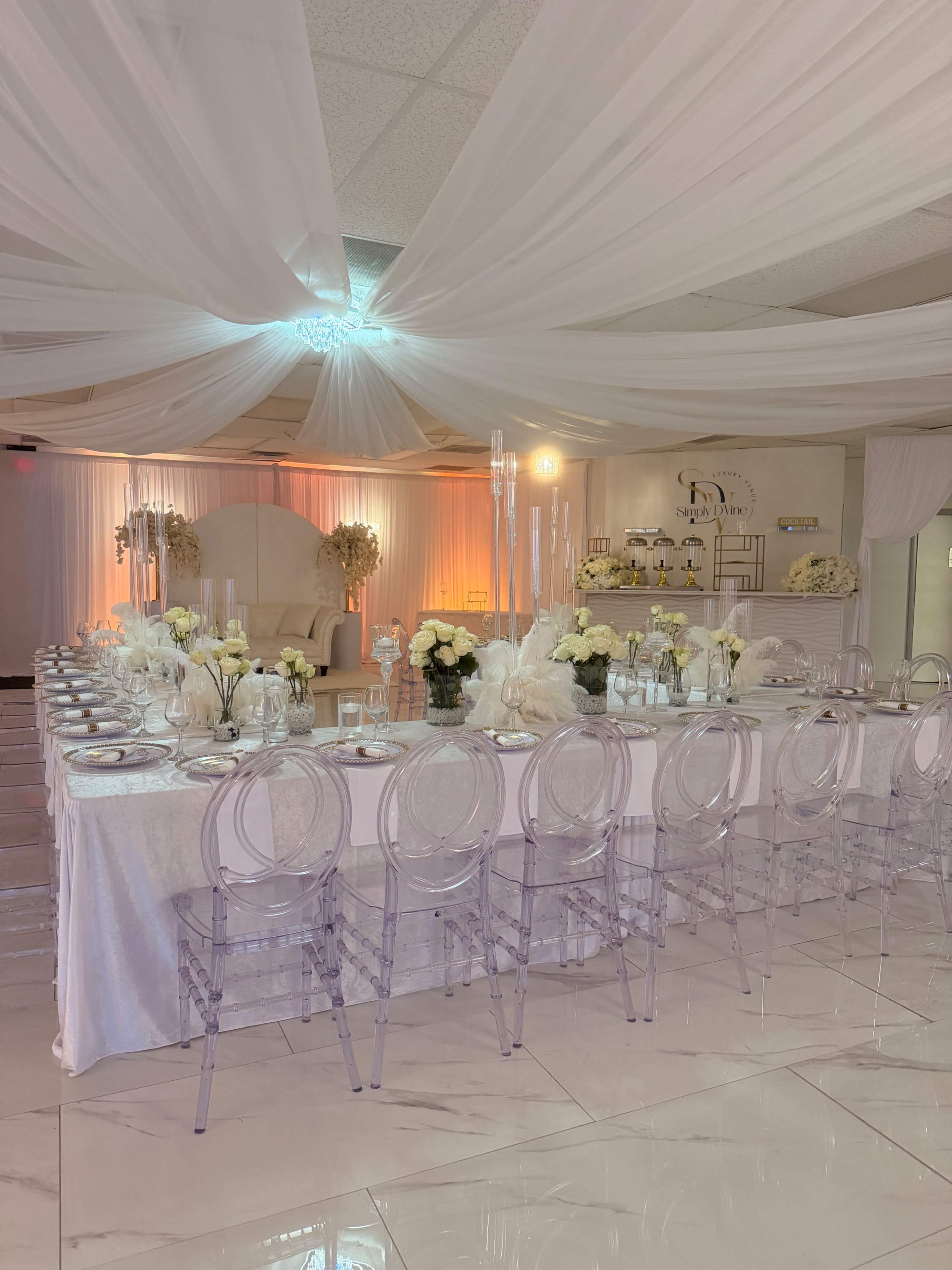 The image shows a beautifully arranged banquet hall with a long dining table set with white tableware and floral centerpieces, surrounded by transparent chairs under draped white fabric.