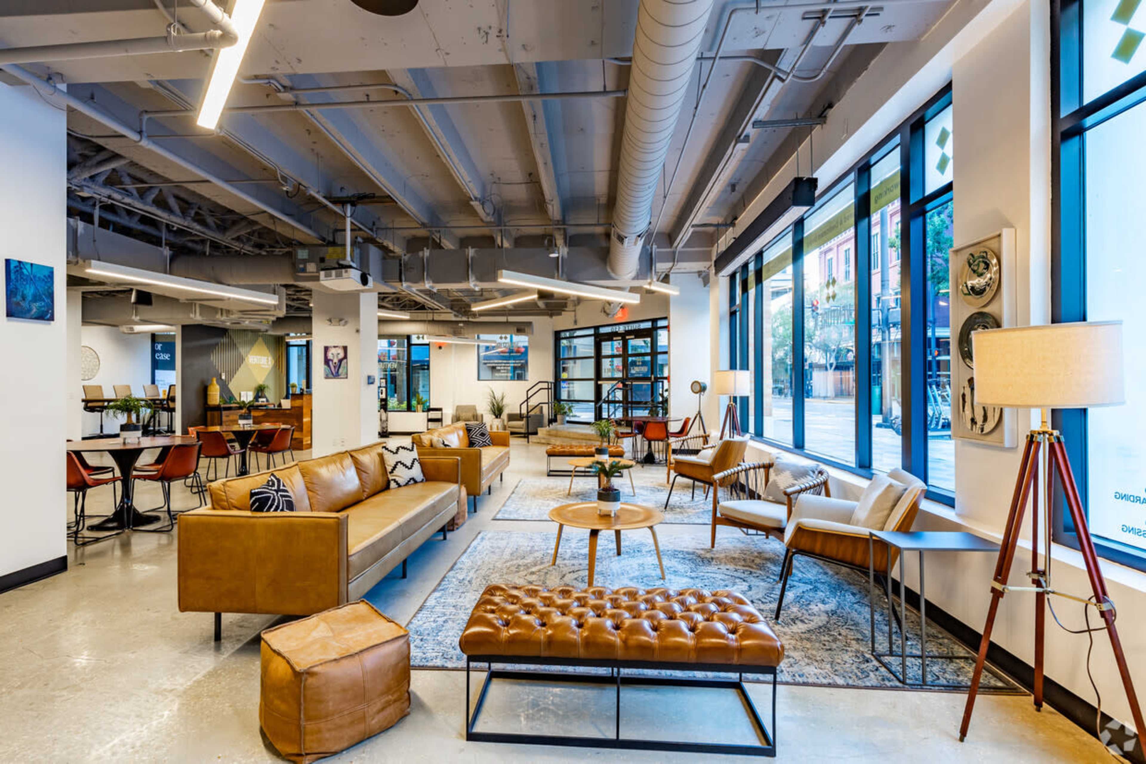 A modern coworking space featuring leather seating, a coffee table, and large windows allowing natural light to enter.