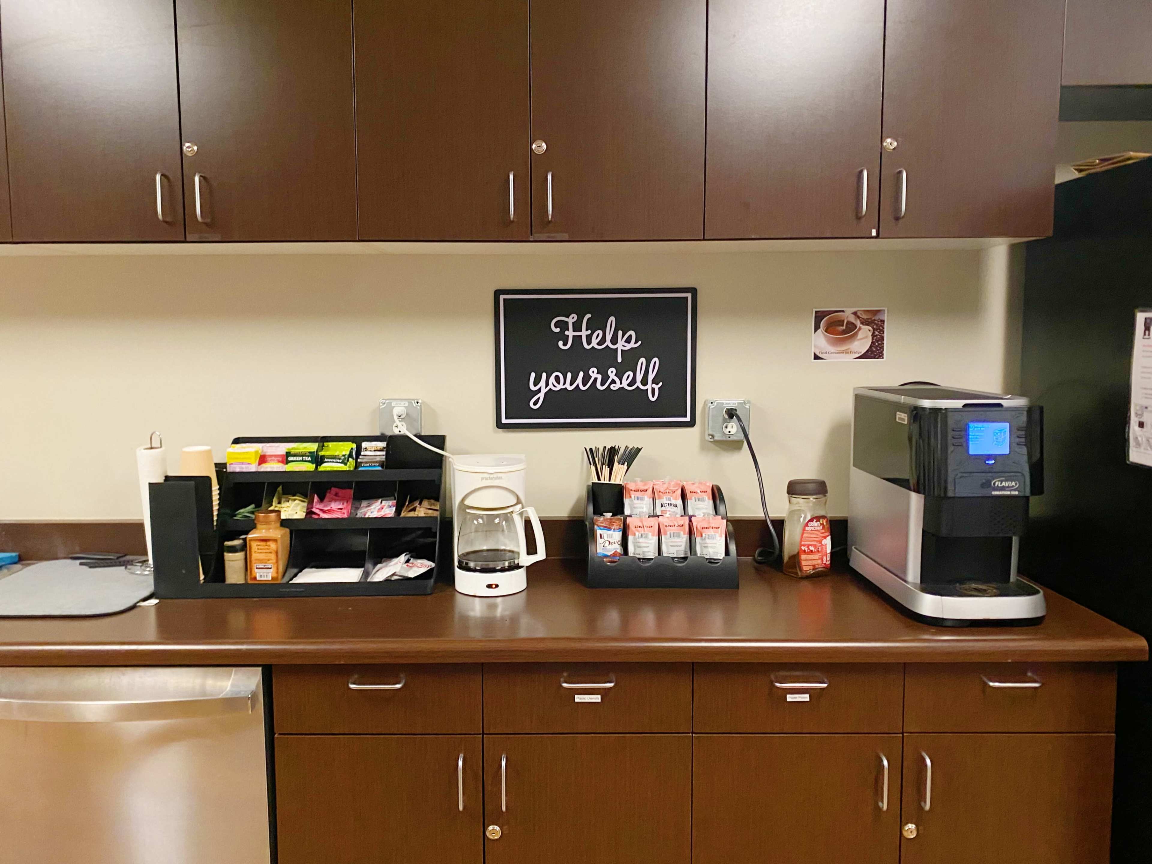 The image depicts a kitchenette with a coffee machine, a variety of tea bags, condiments, and supplies organized on a countertop beneath a sign that says "Help yourself."