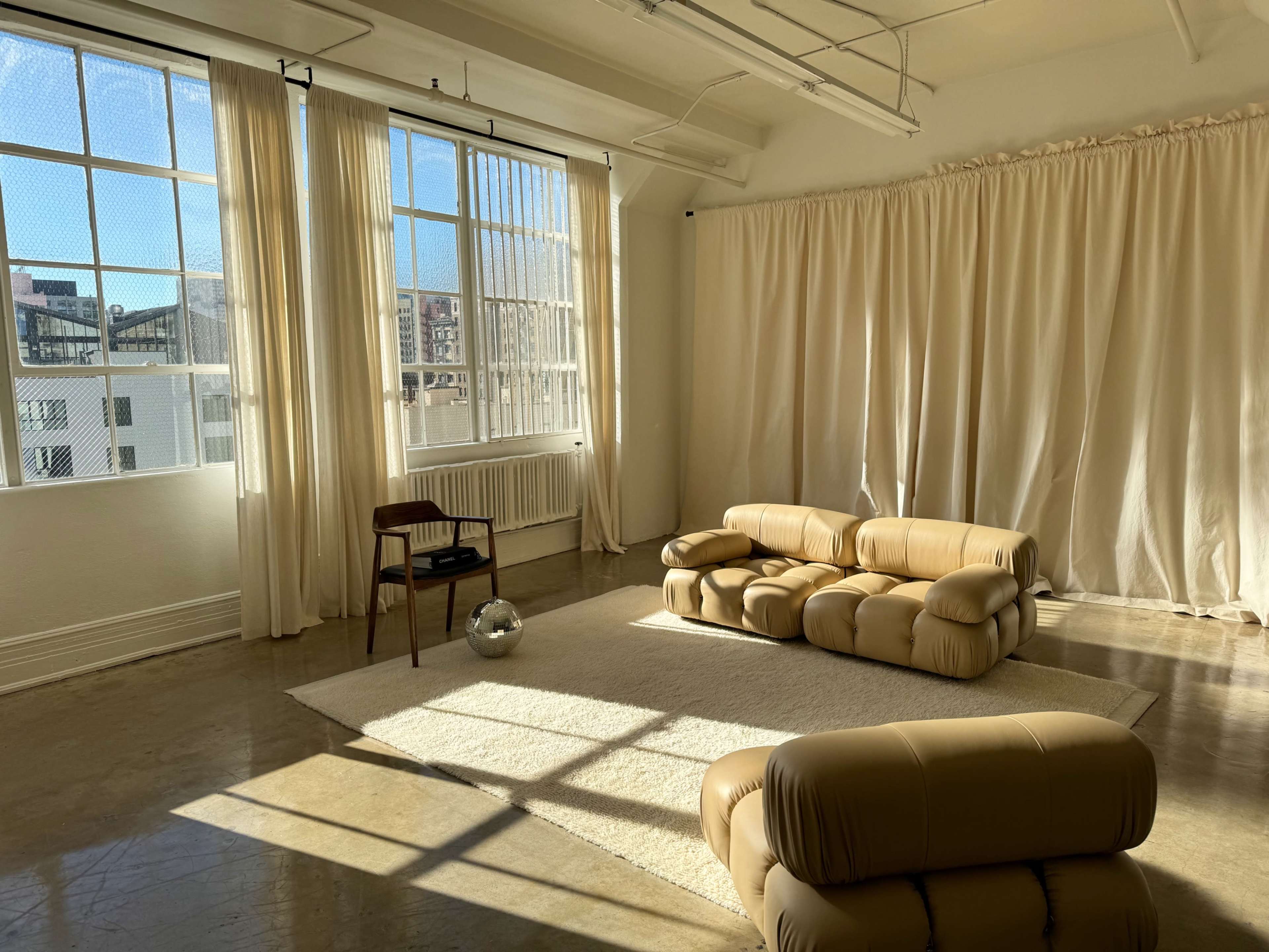 A spacious room with large windows allowing natural light, a textured rug on the floor, a minimalist lounge chair, and a sofa arranged near white curtains.