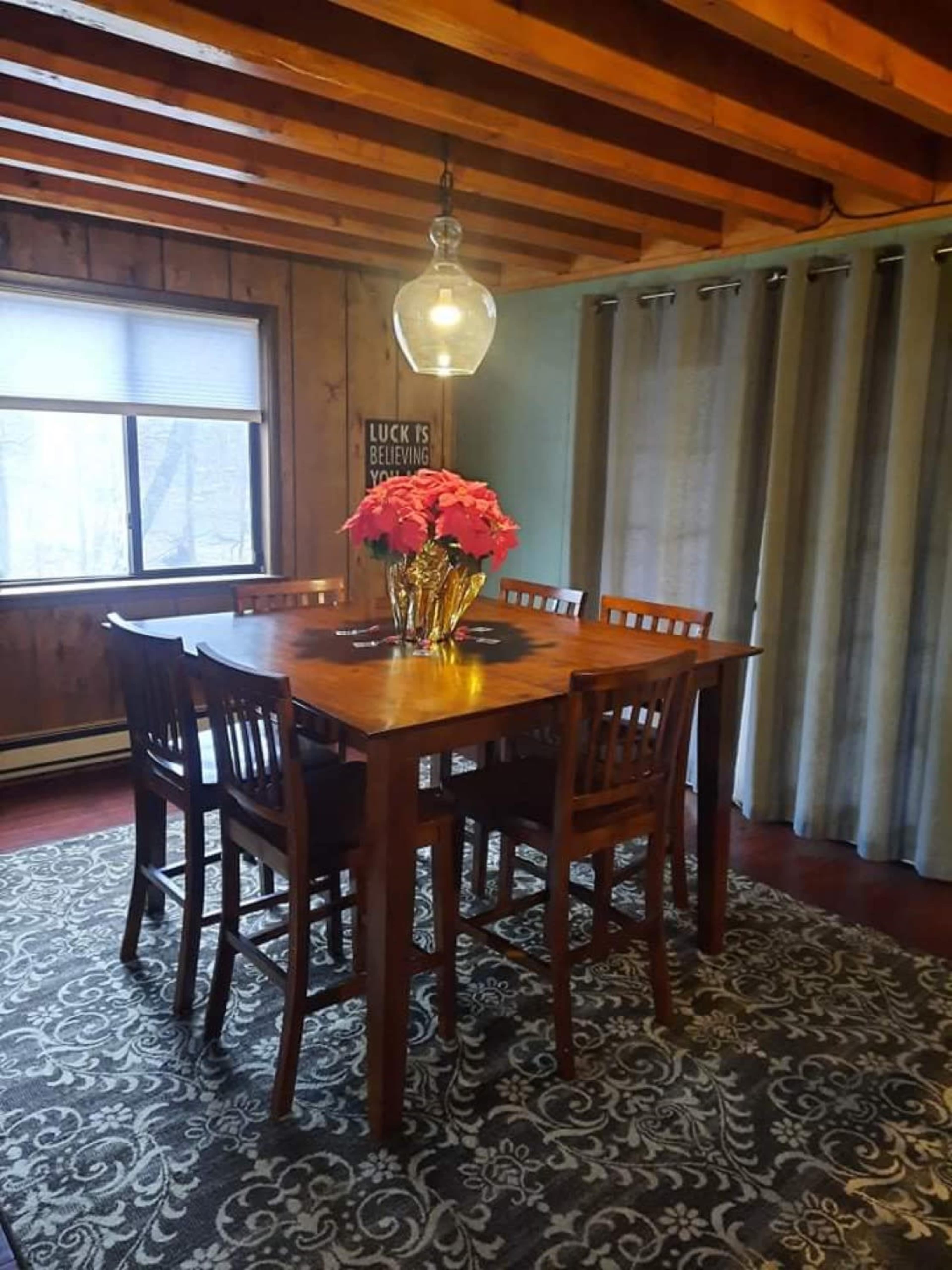 A wooden dining table surrounded by high-backed chairs is set under a pendant light in a room with wooden walls and a large window.
