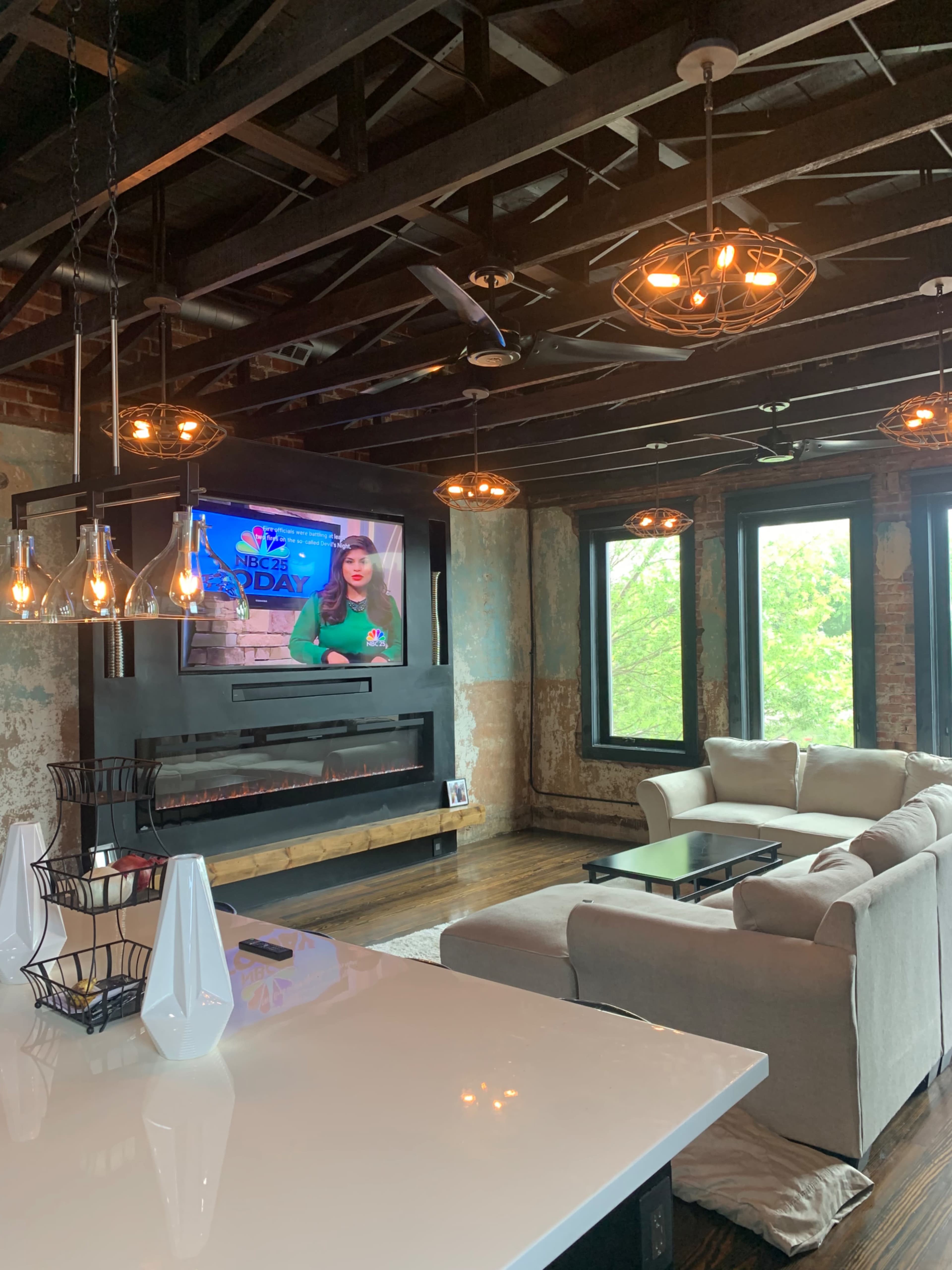 A modern living room features a large flat-screen TV mounted on a wall above a sleek fireplace, with a sectional sofa and several windows allowing natural light to enter.