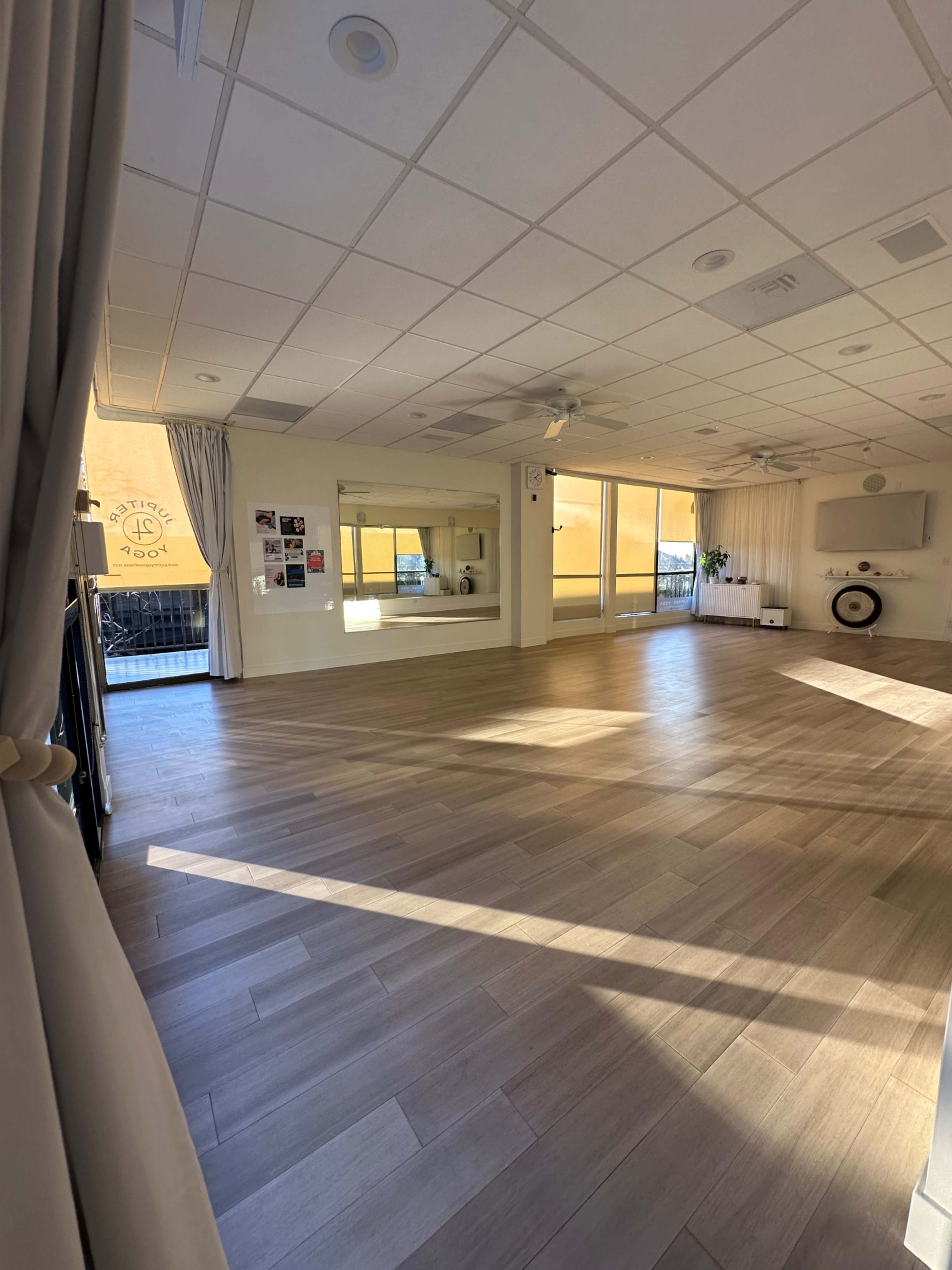 The image shows a spacious, well-lit room with wooden floors, large windows, and mirrors along one wall.