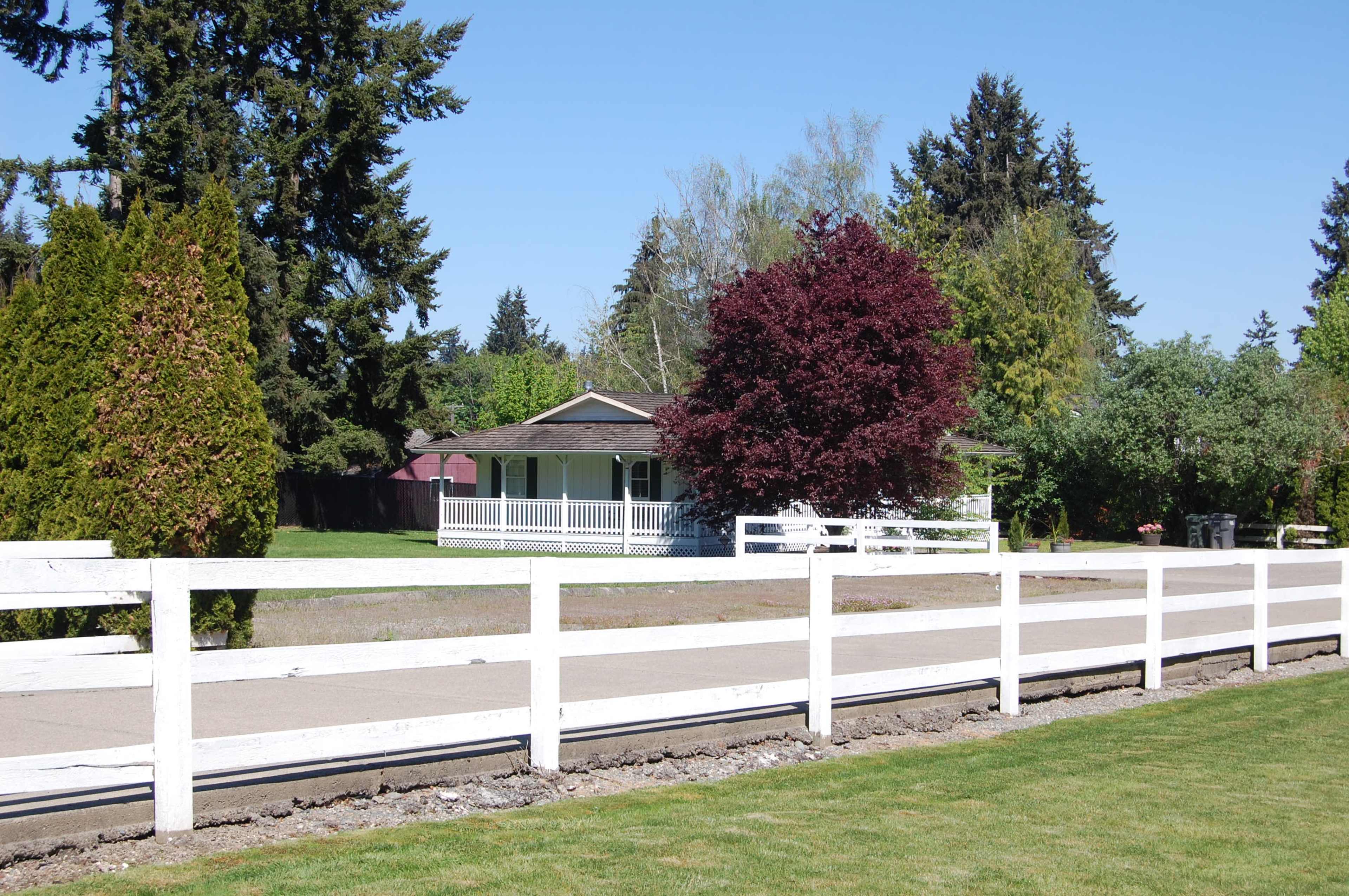 A single-story house with a wraparound porch is surrounded by greenery, with a white wooden fence in the foreground.