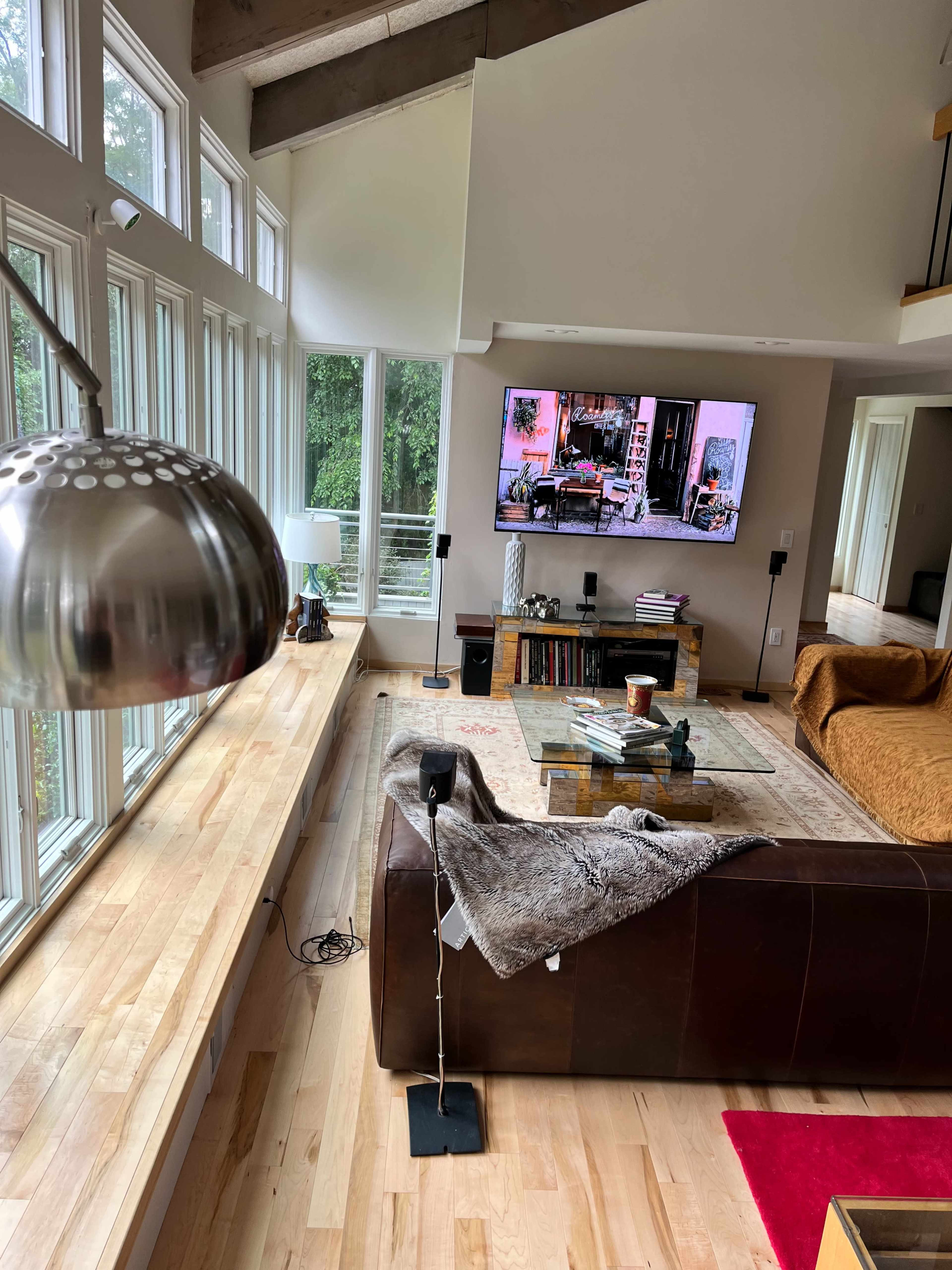 The image shows a spacious living room with large windows, a modern television displaying a scene, a brown leather couch, and wooden flooring.