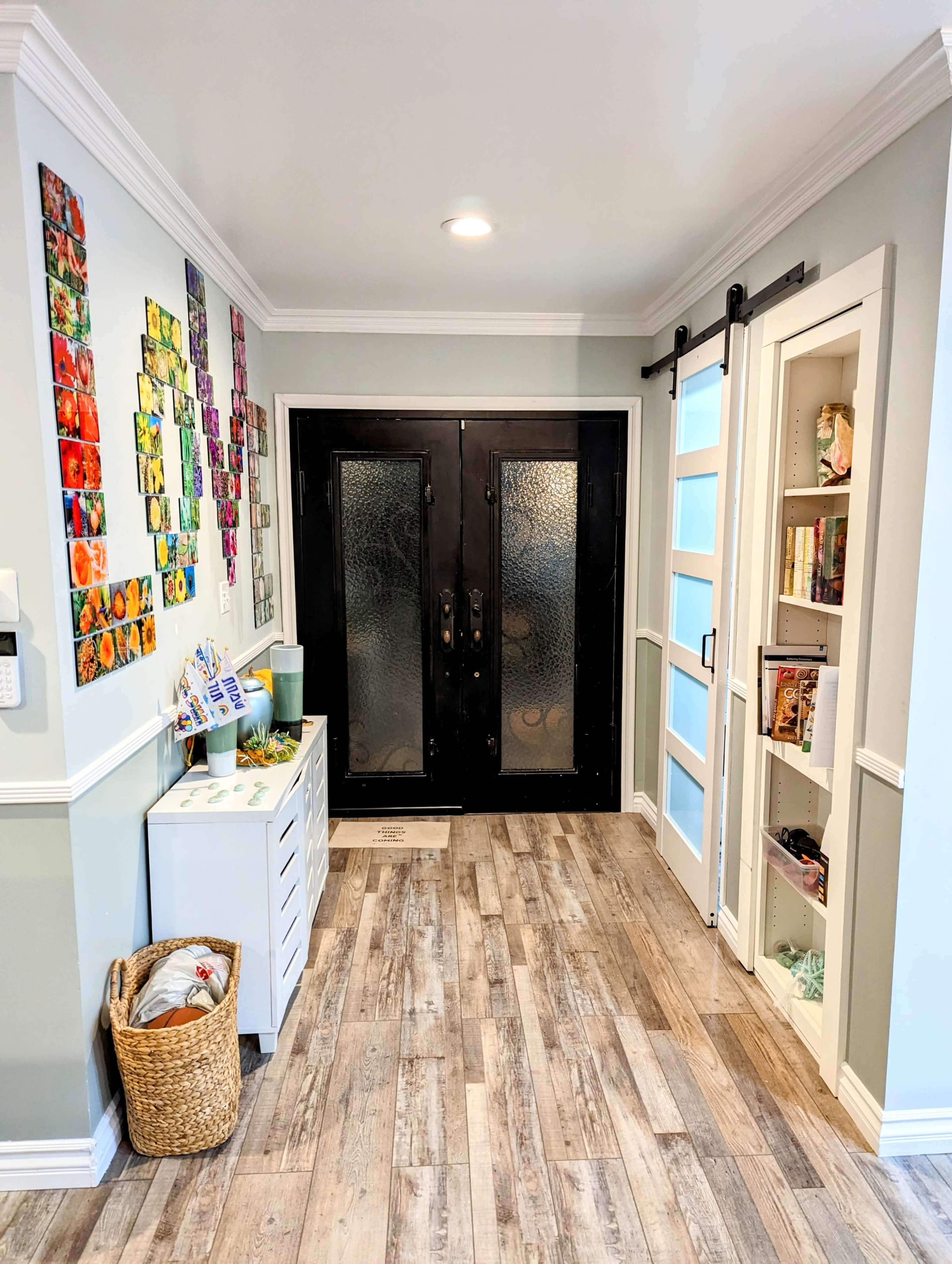 The image shows a well-lit entryway with black double doors, a decorated wall of colorful artwork, and a small storage cabinet beside a shelf.