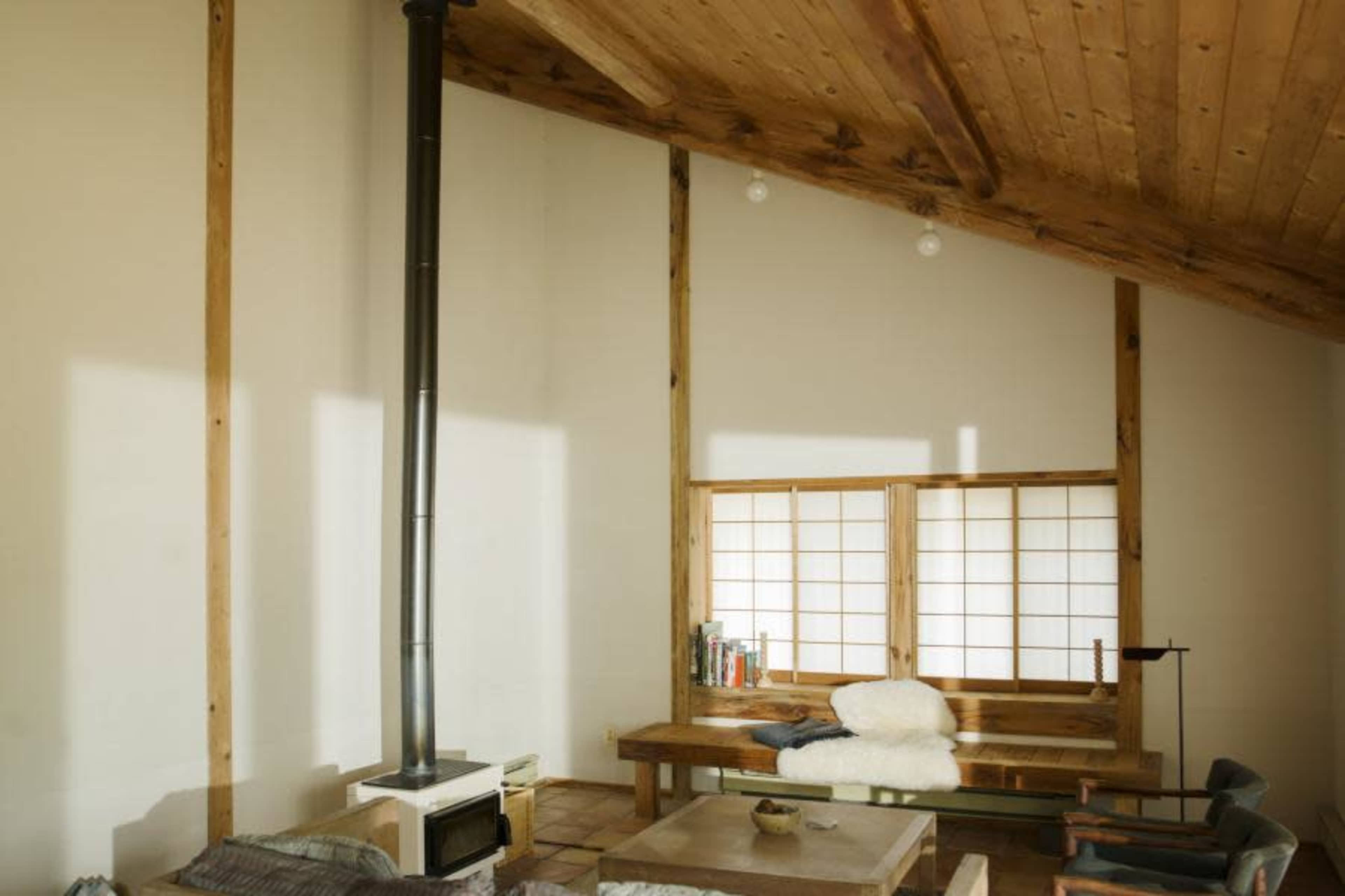 The image shows a wooden interior with a high ceiling, featuring a stove, a bench with a fur throw, and a table on a wooden floor.