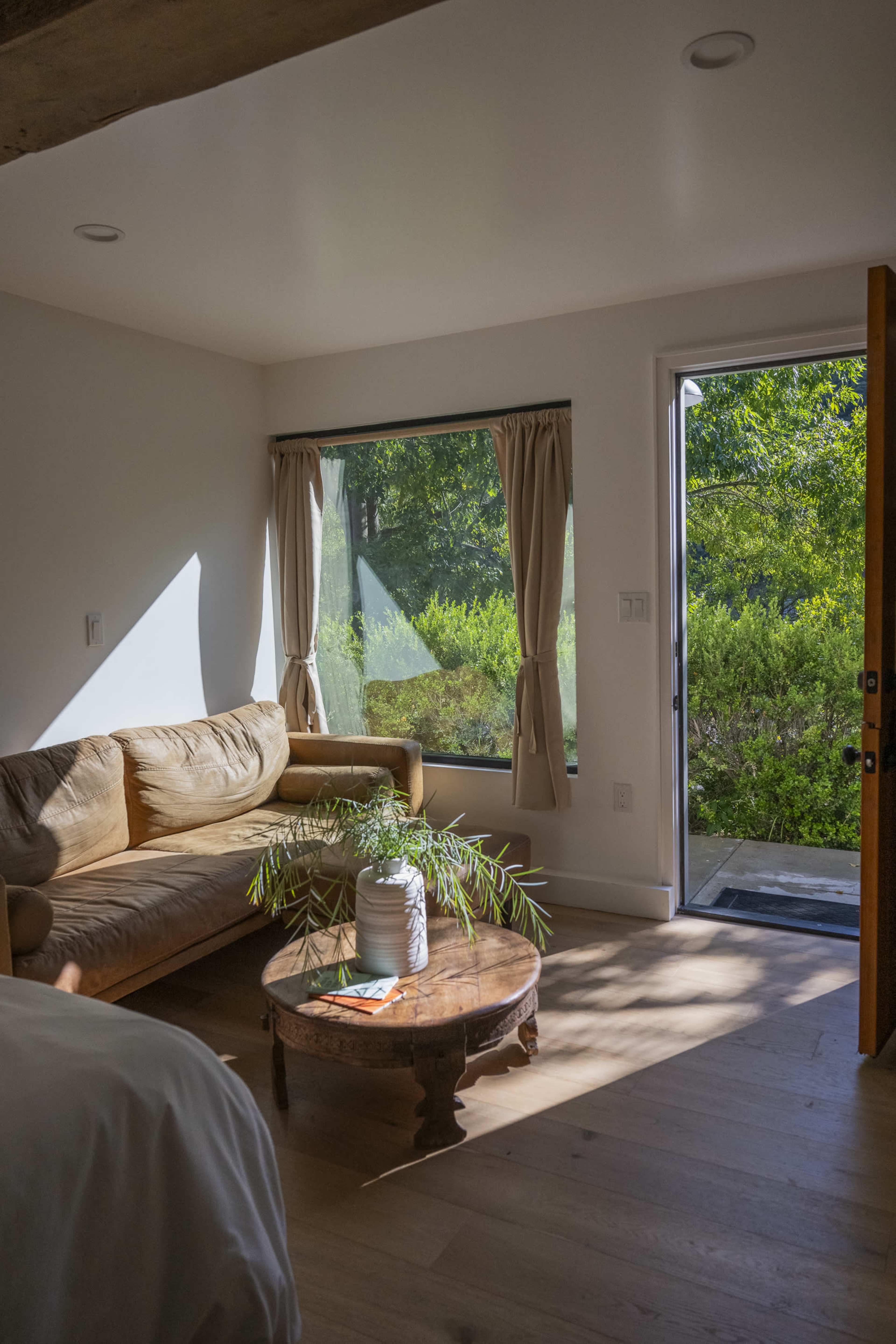 The image shows a cozy bedroom with a sofa, a wooden coffee table, and a doorway leading to an outdoor view of greenery.