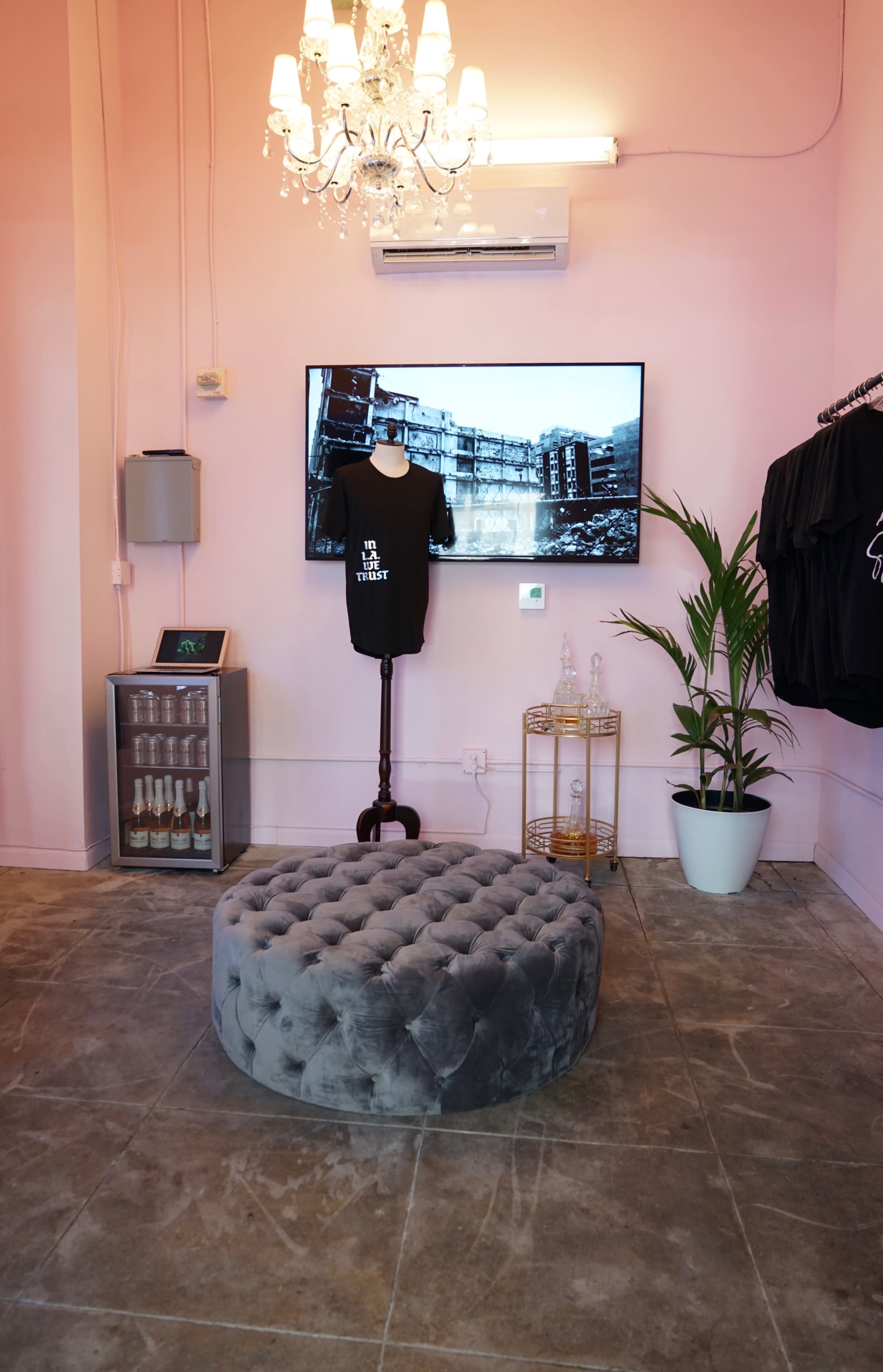 A pink-walled boutique featuring a tufted gray ottoman, a wall-mounted television, a plant in a white pot, and clothing displayed on hangers.