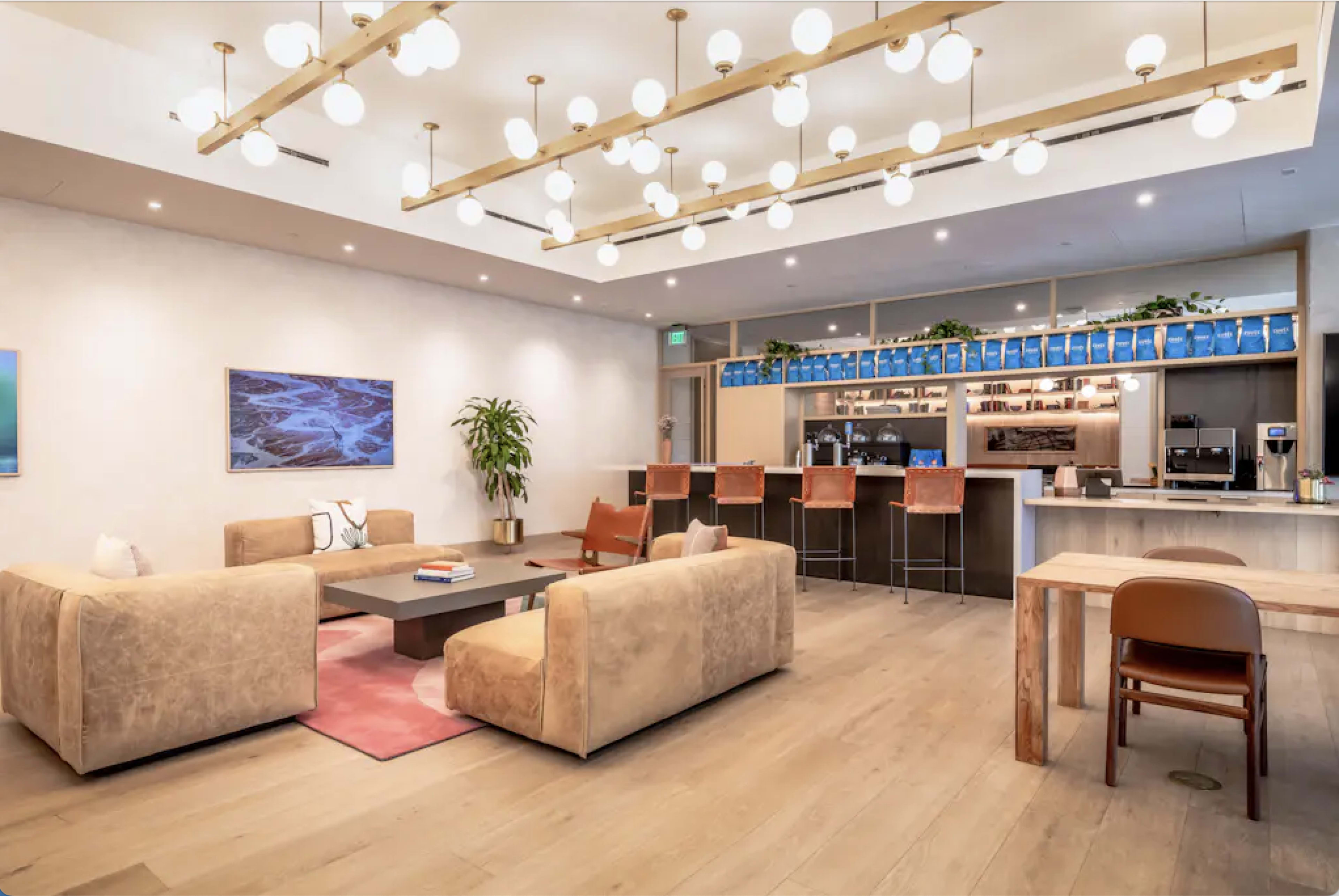The image shows a modern lounge area with two beige sofas, a coffee table in the center, a wooden dining table, and a bar counter with stools illuminated by decorative lighting.