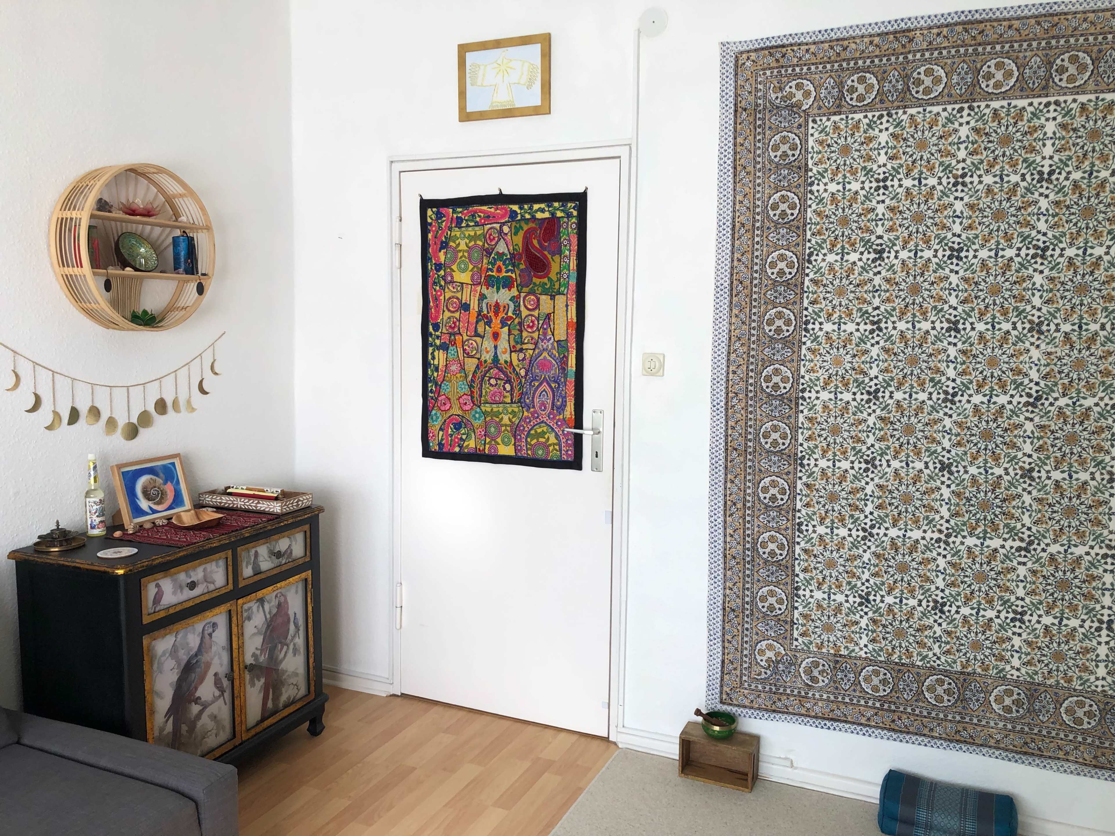A bright room featuring a doorway with a decorative fabric hanging, a patterned wall tapestry, a wooden cabinet with framed photos, and a gray sofa.