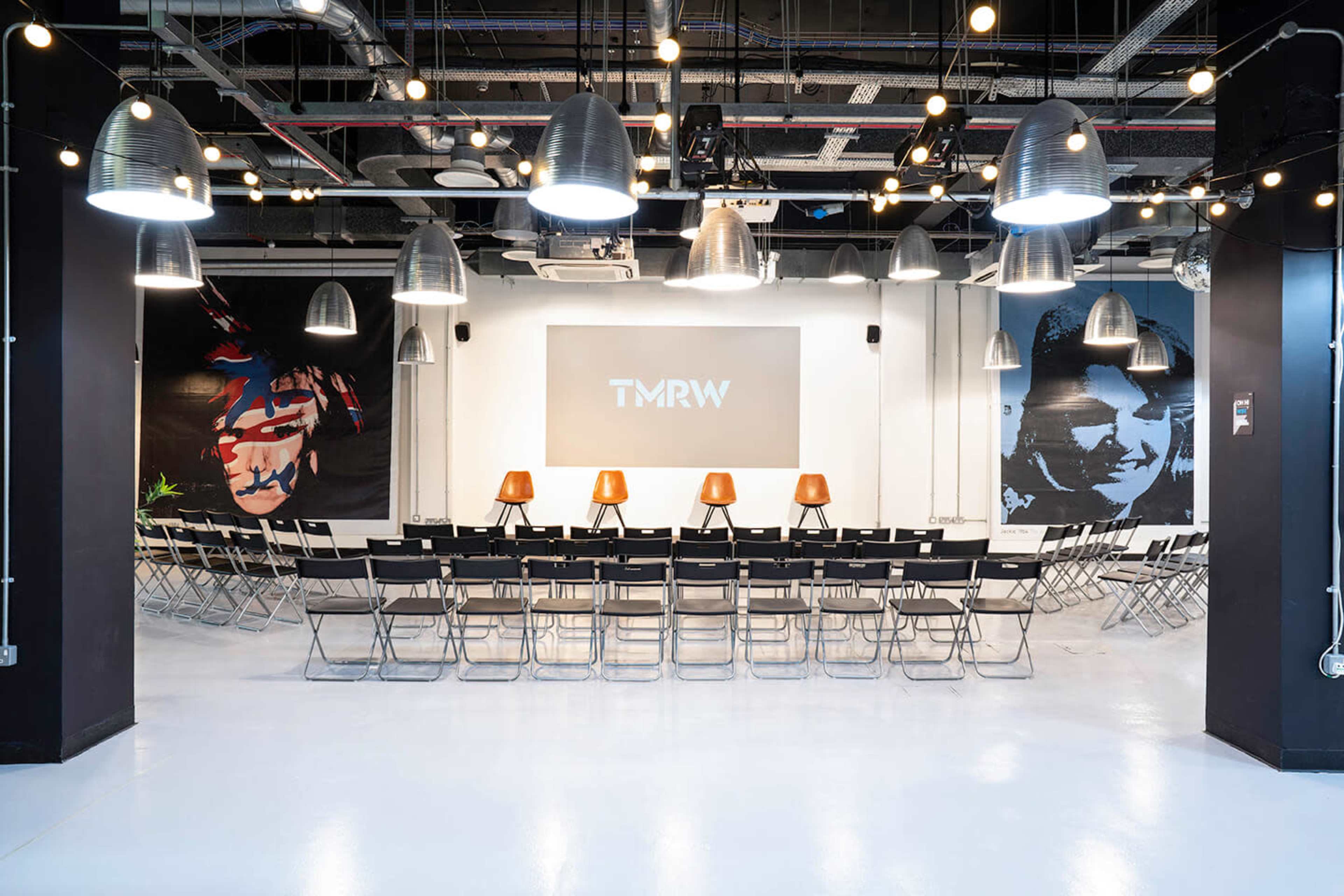 A modern event space with rows of black folding chairs facing a large screen, illuminated by pendant lights and featuring murals on the walls.