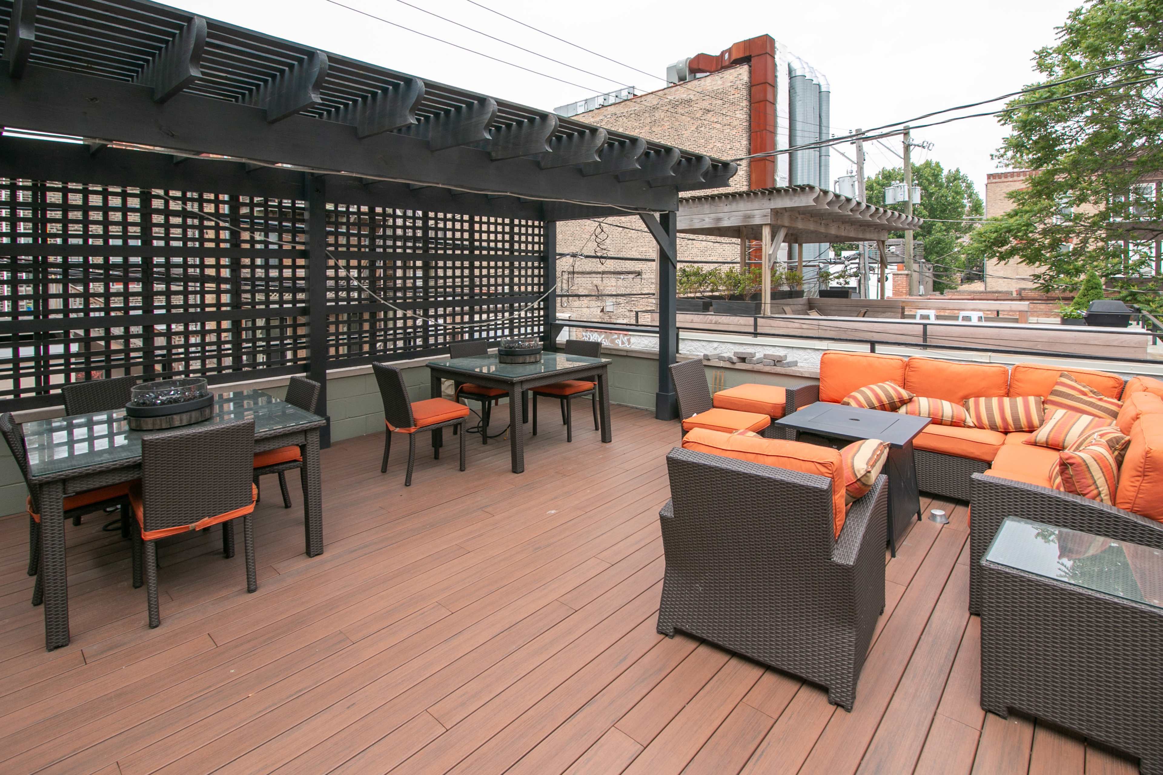 The image shows a rooftop patio featuring orange seating, black wicker furniture, and several tables arranged on a wooden deck.