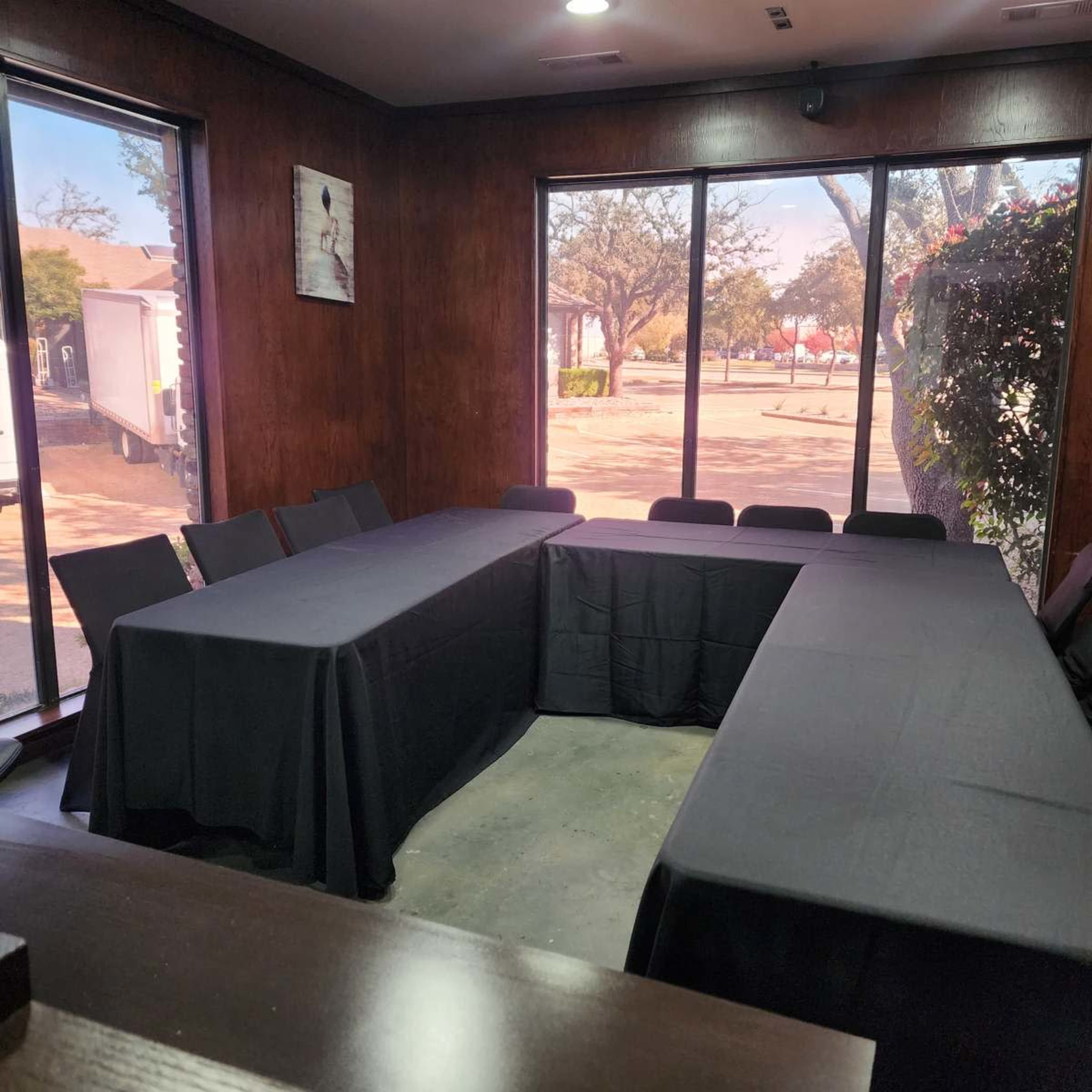 A meeting room with black tablecloths covering two rectangular tables arranged in a U-shape, surrounded by black chairs and large windows letting in natural light.