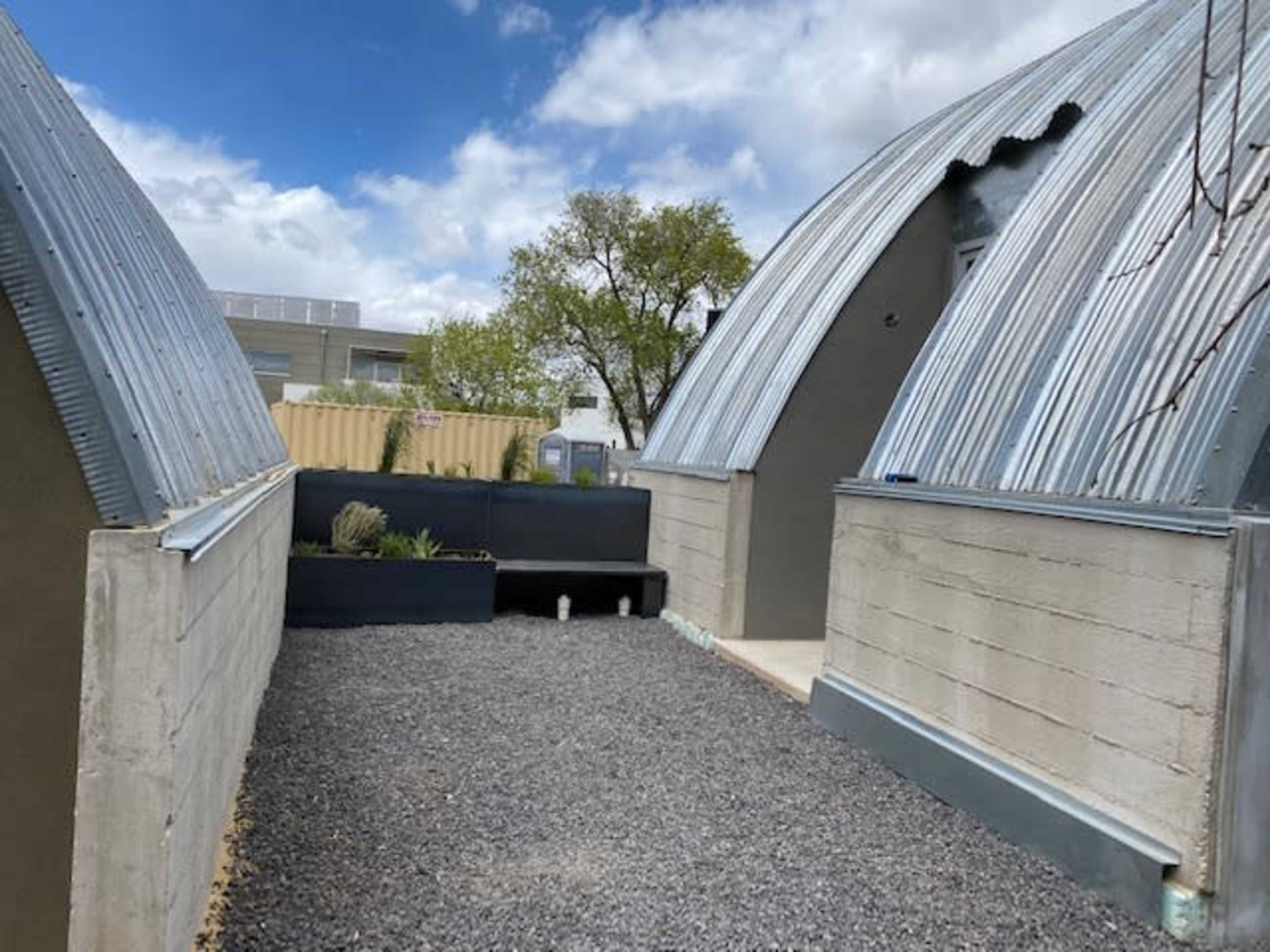 The image shows a courtyard between two curved metal-roofed structures, with gravel on the ground and some plants in black containers.