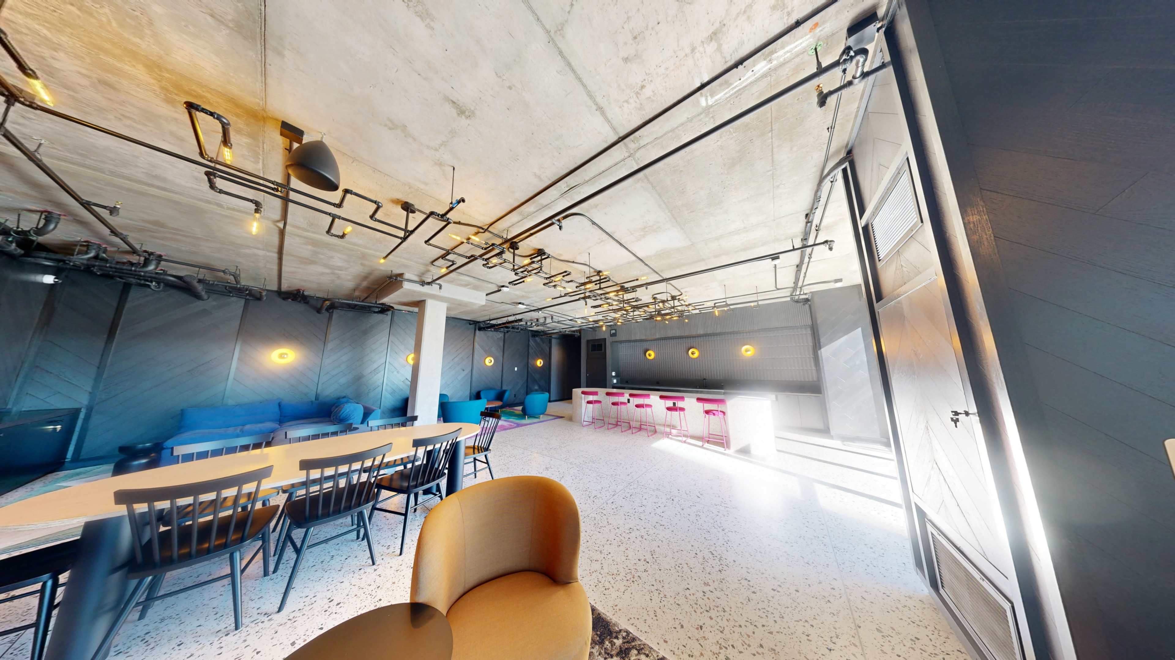The image shows a modern, industrial-style interior space with a long table, seating areas, and a bar counter illuminated by overhead lighting.