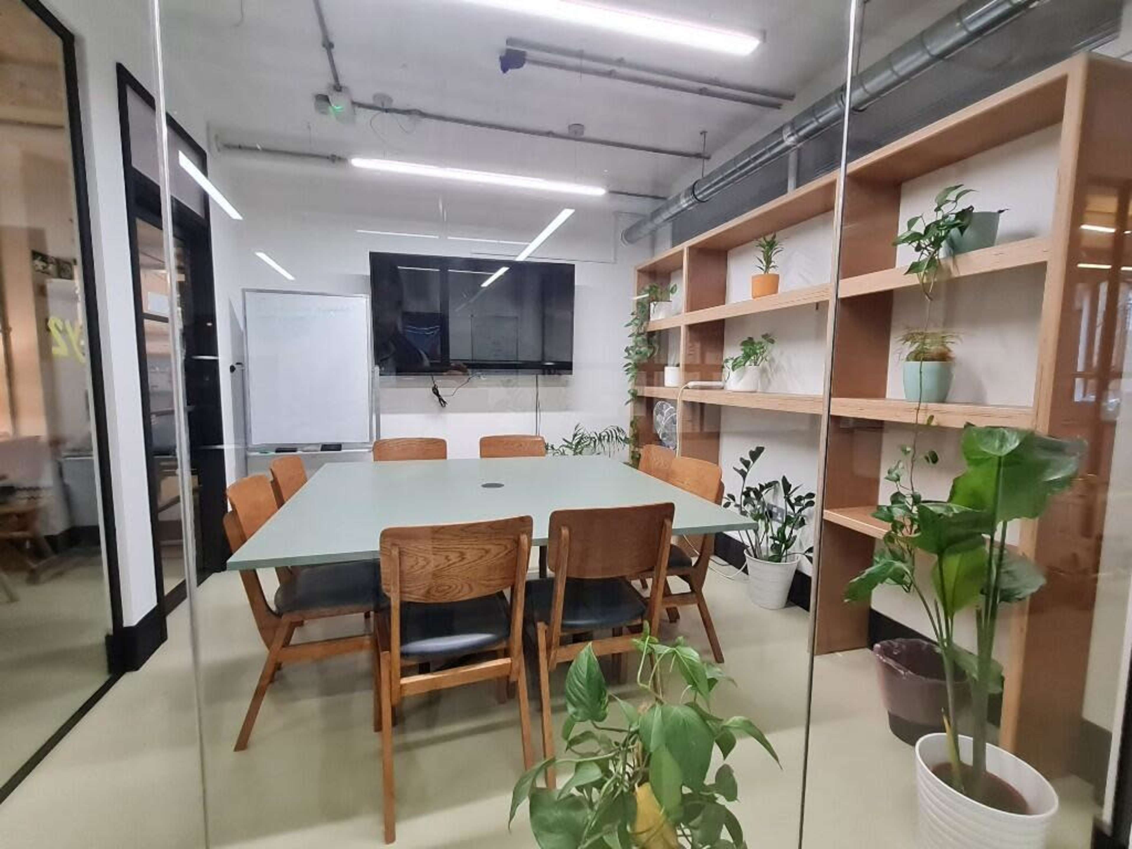 The image shows a modern conference room featuring a rectangular table surrounded by wooden chairs, with plants and a wall-mounted TV in the background.