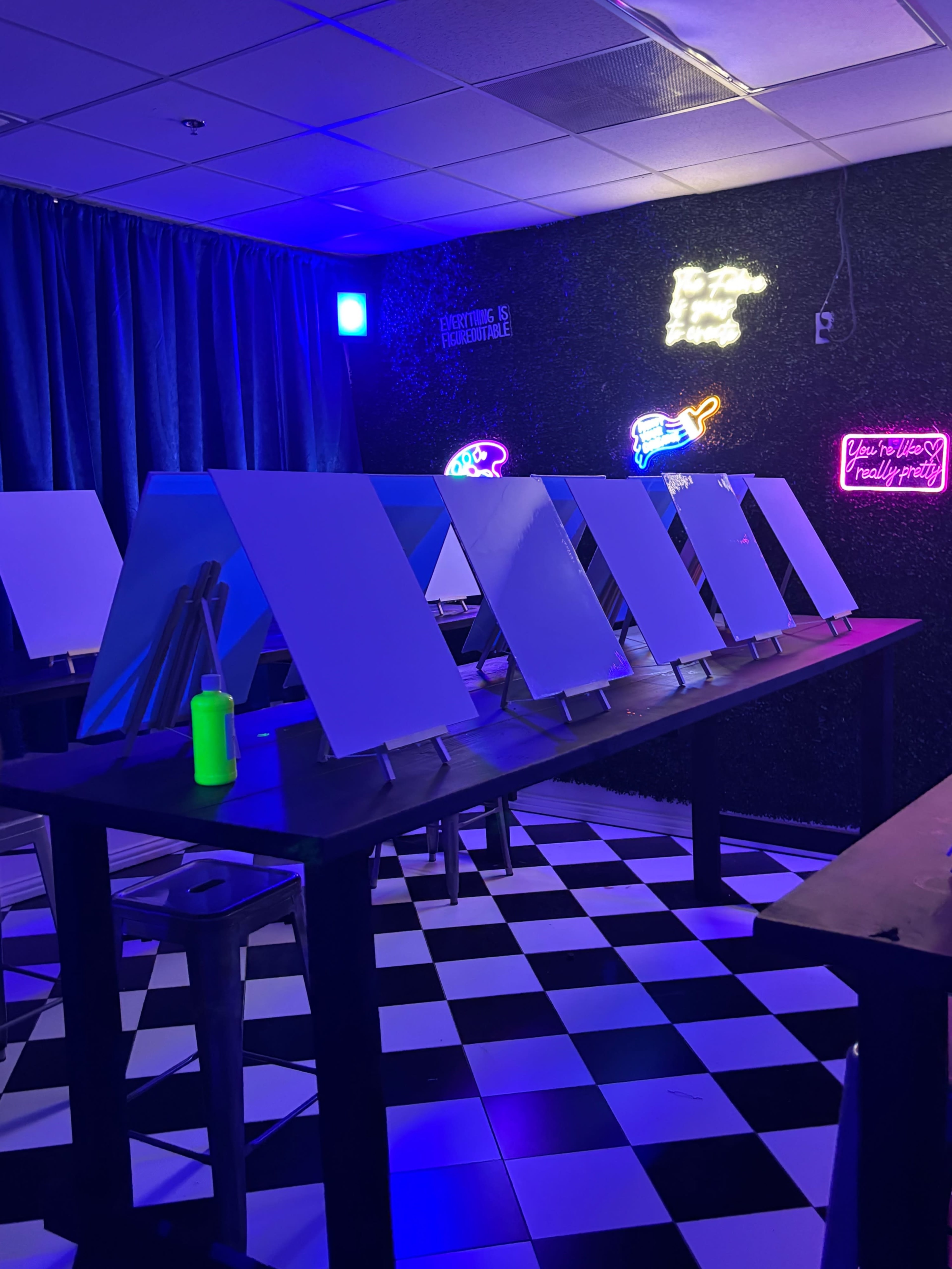 The image shows a spacious art studio with several blank easels positioned on tables, illuminated by colorful neon signs and ambient lighting.