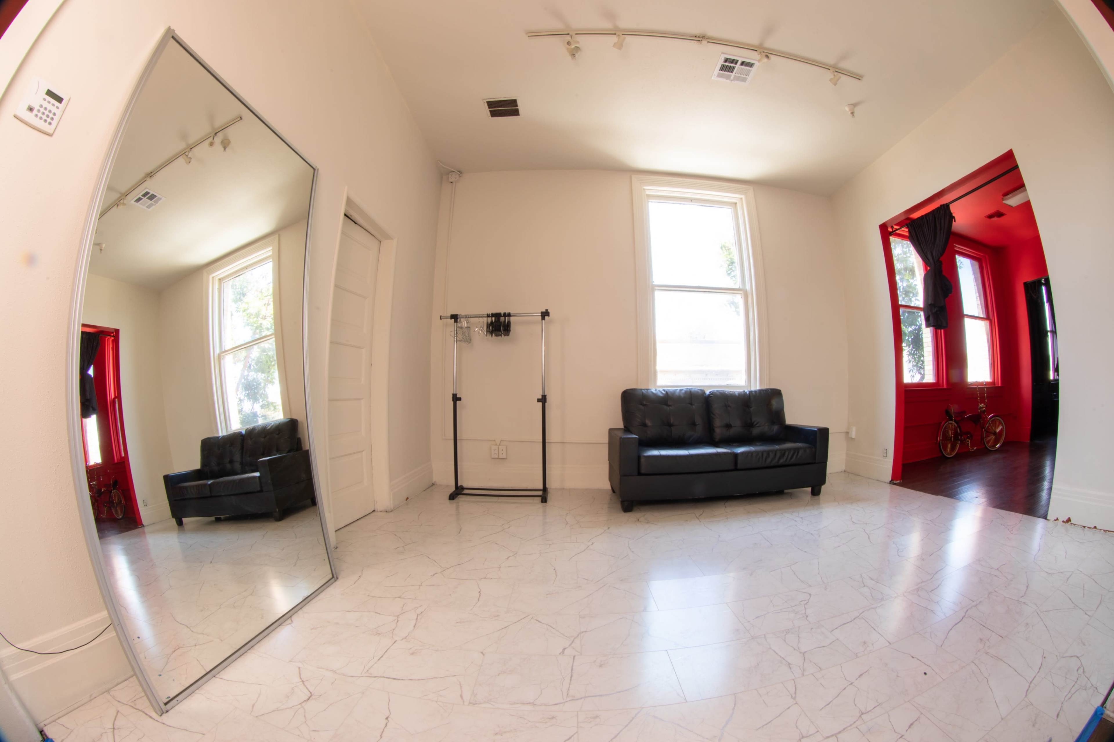 The image shows a spacious room with a large mirror, a black sofa, and a clothing rack, along with two windows and red accents in the adjacent area.