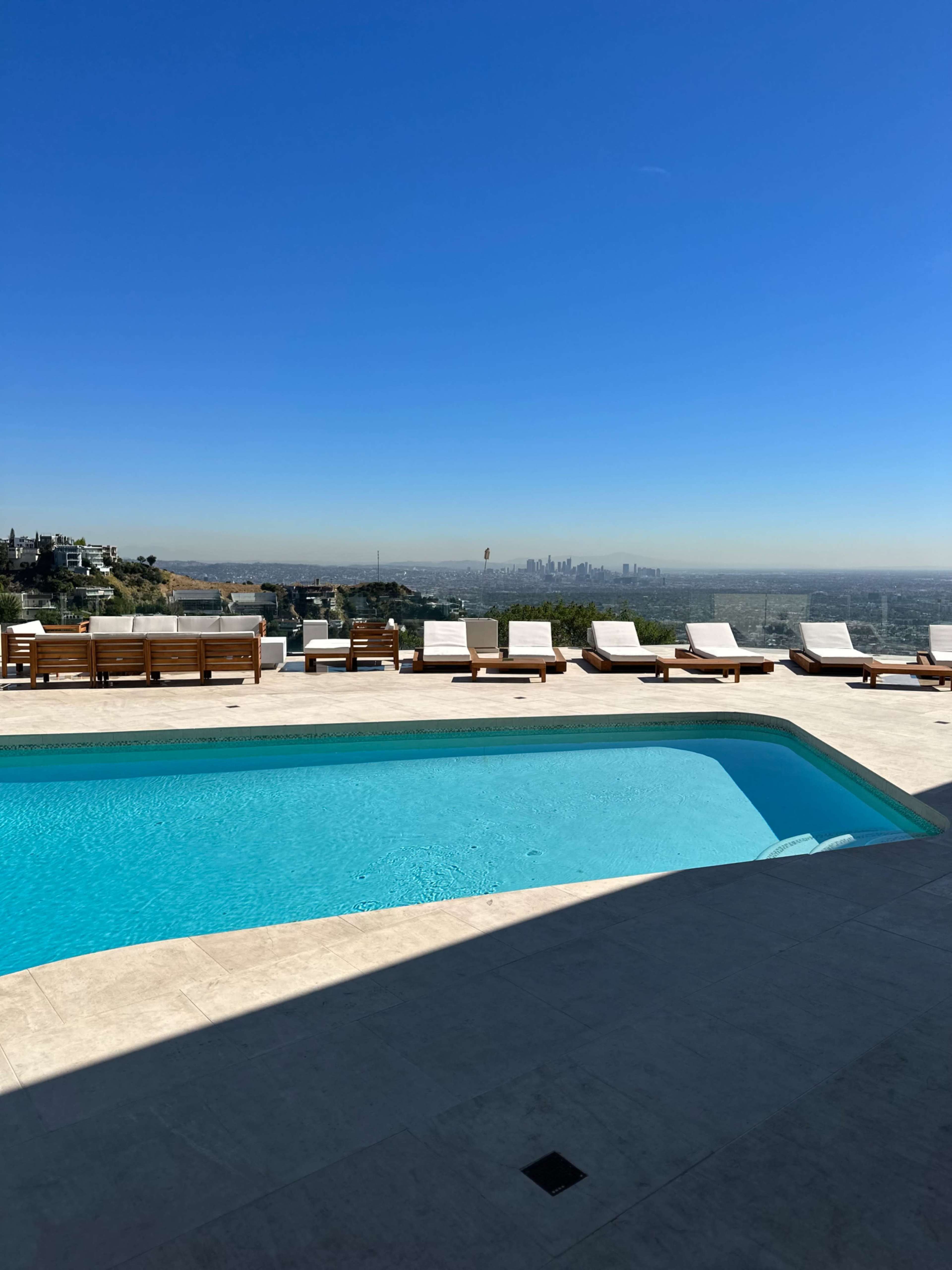 A swimming pool with lounge chairs surrounds it overlooks a city skyline under a clear blue sky.