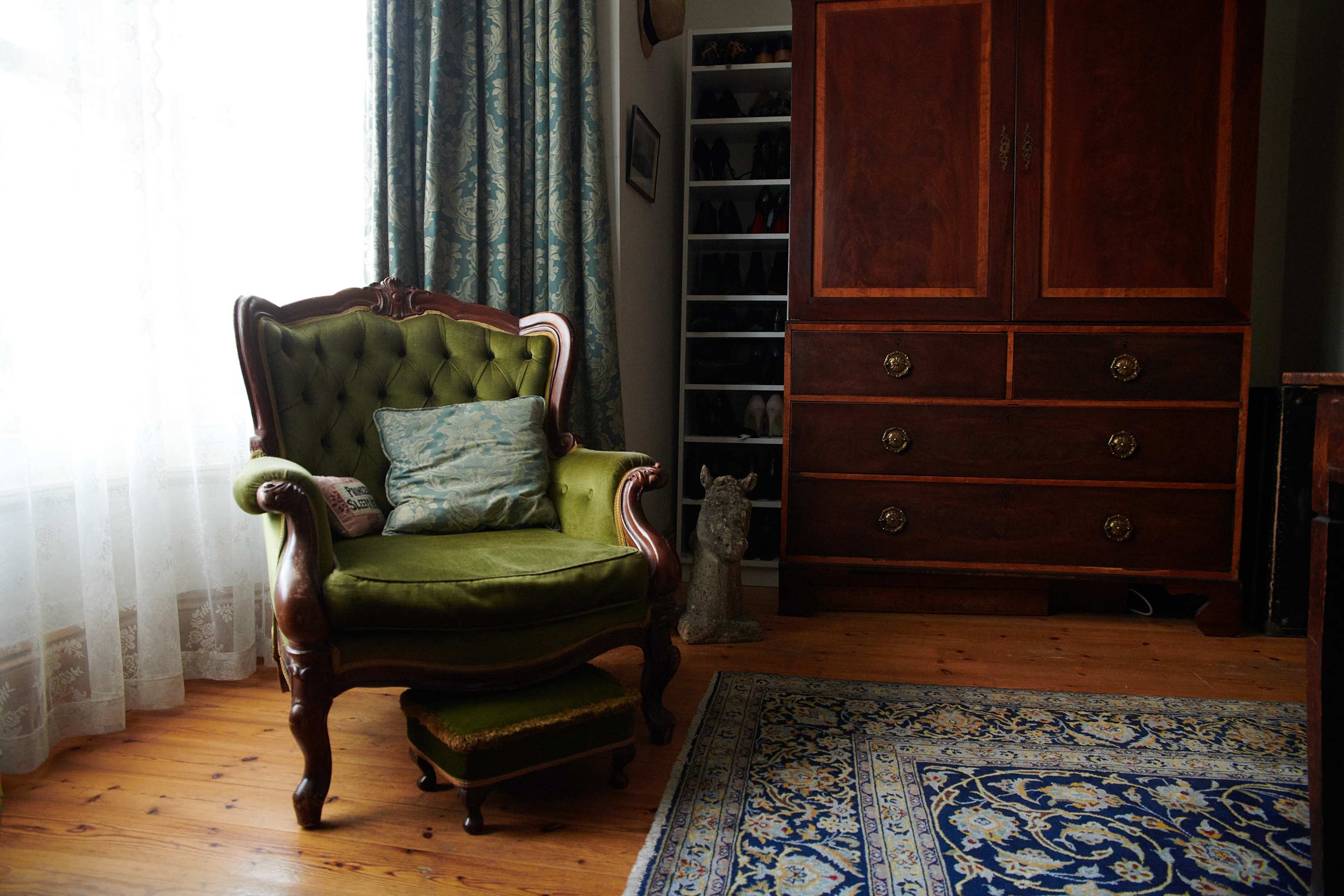 A green upholstered Victorian armchair beside a wooden cabinet and a patterned blue rug on a wooden floor.