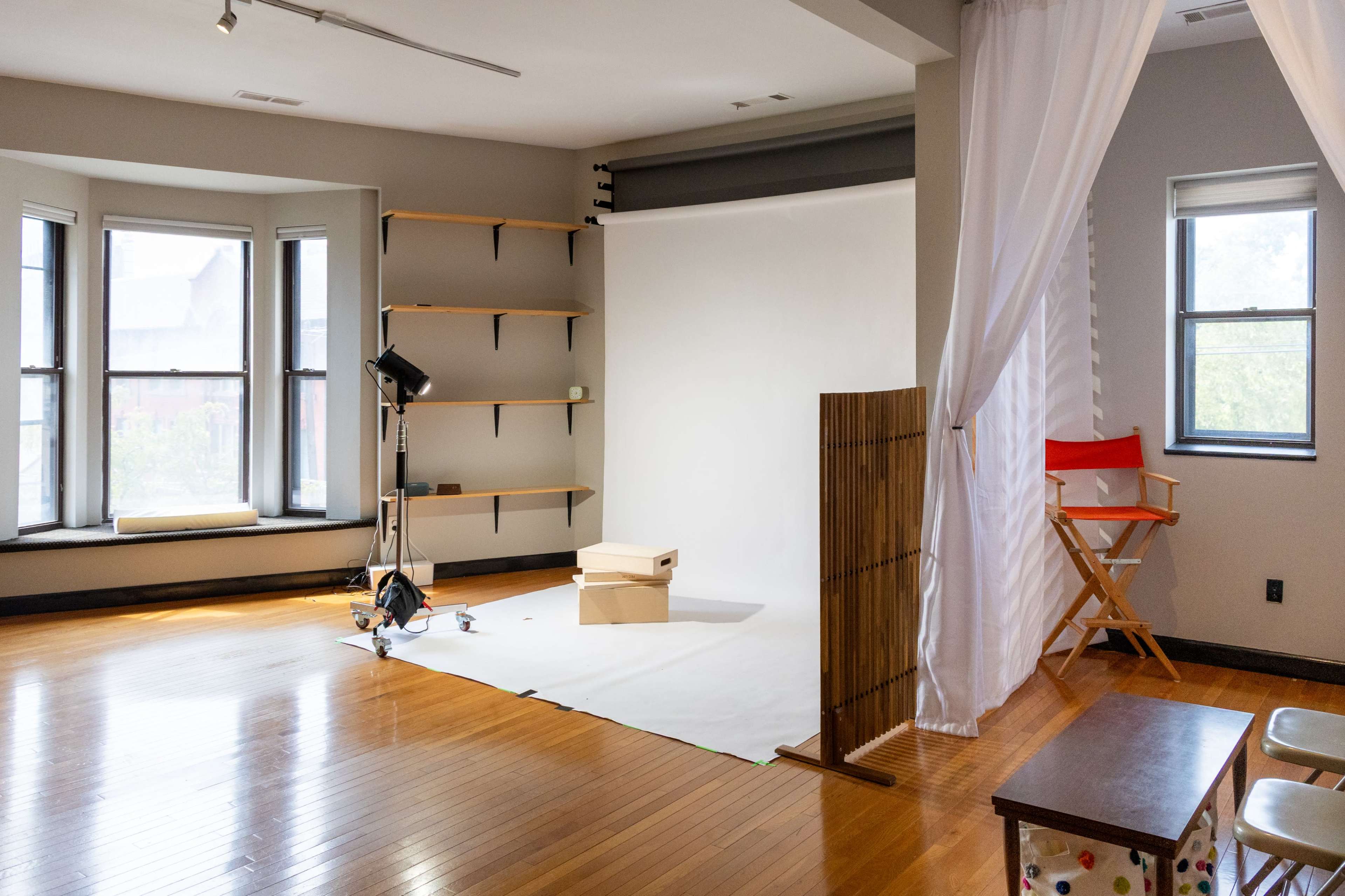 The image shows a photography studio with an empty backdrop, two windows, wooden shelves, a light source, and a red director's chair.