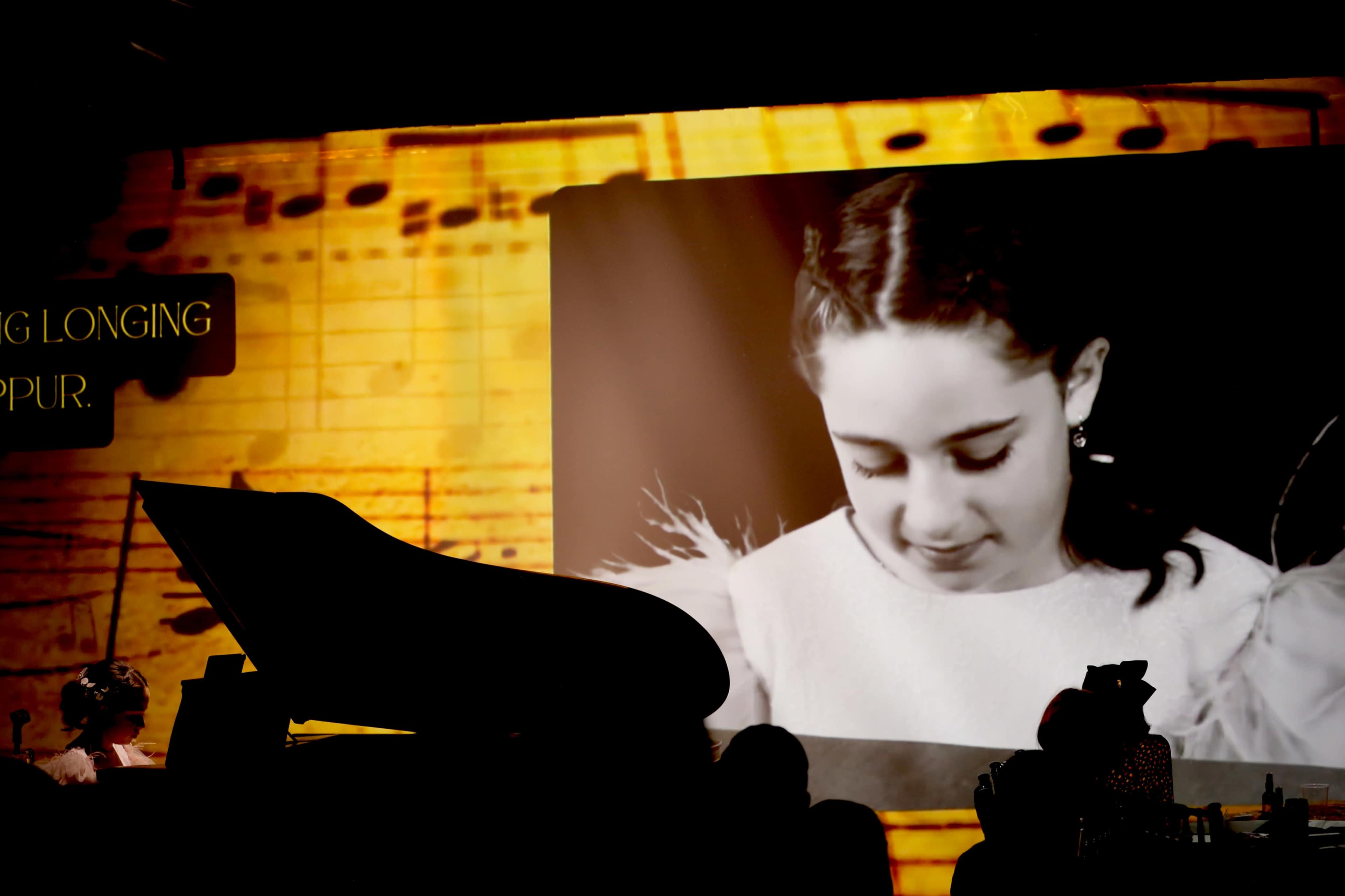 A silhouetted figure sits at a piano in front of a large screen displaying a black-and-white image of a young girl.
