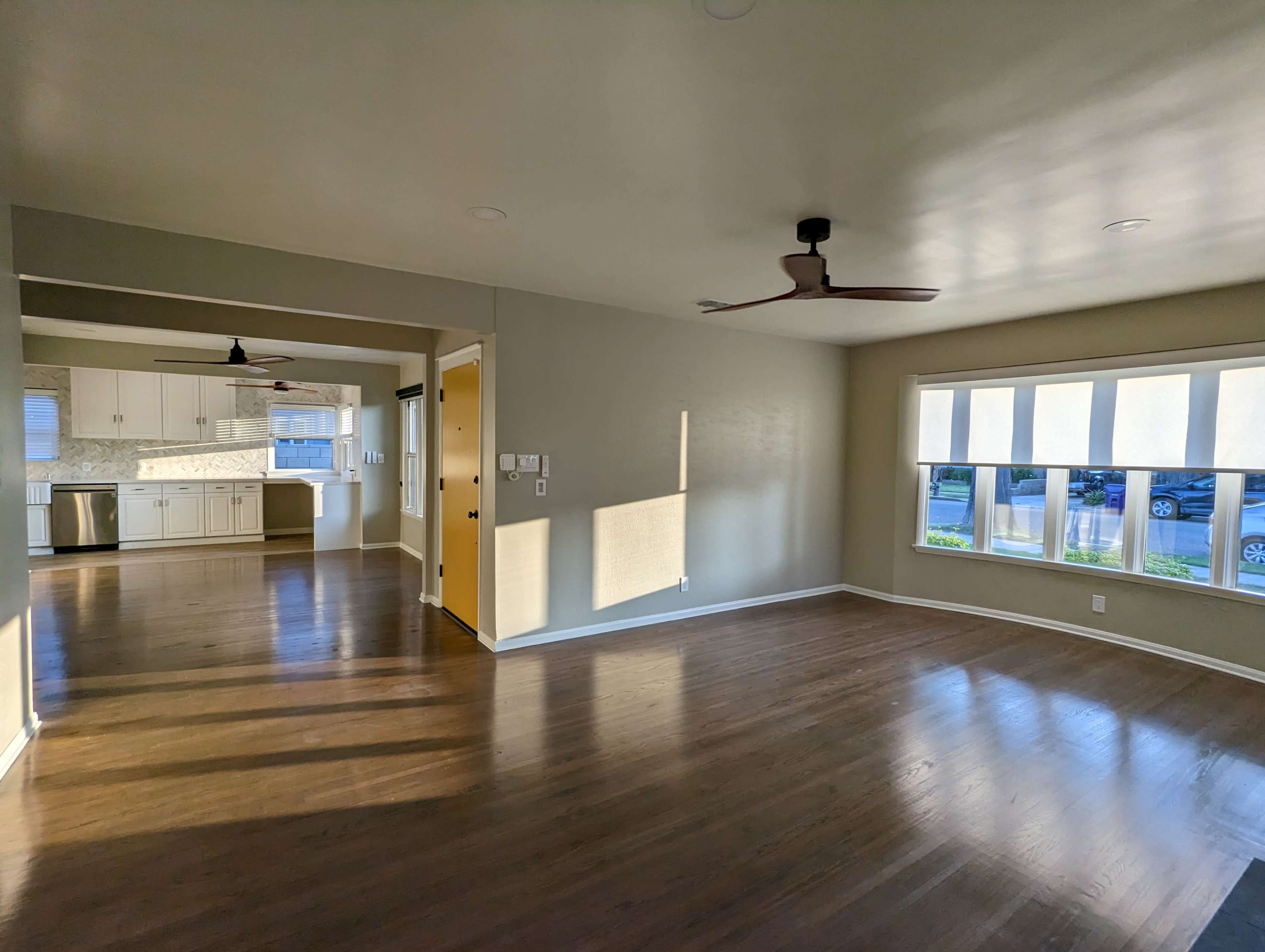 The image shows a spacious interior room with hardwood floors, large windows, and an open layout leading into a kitchen area.