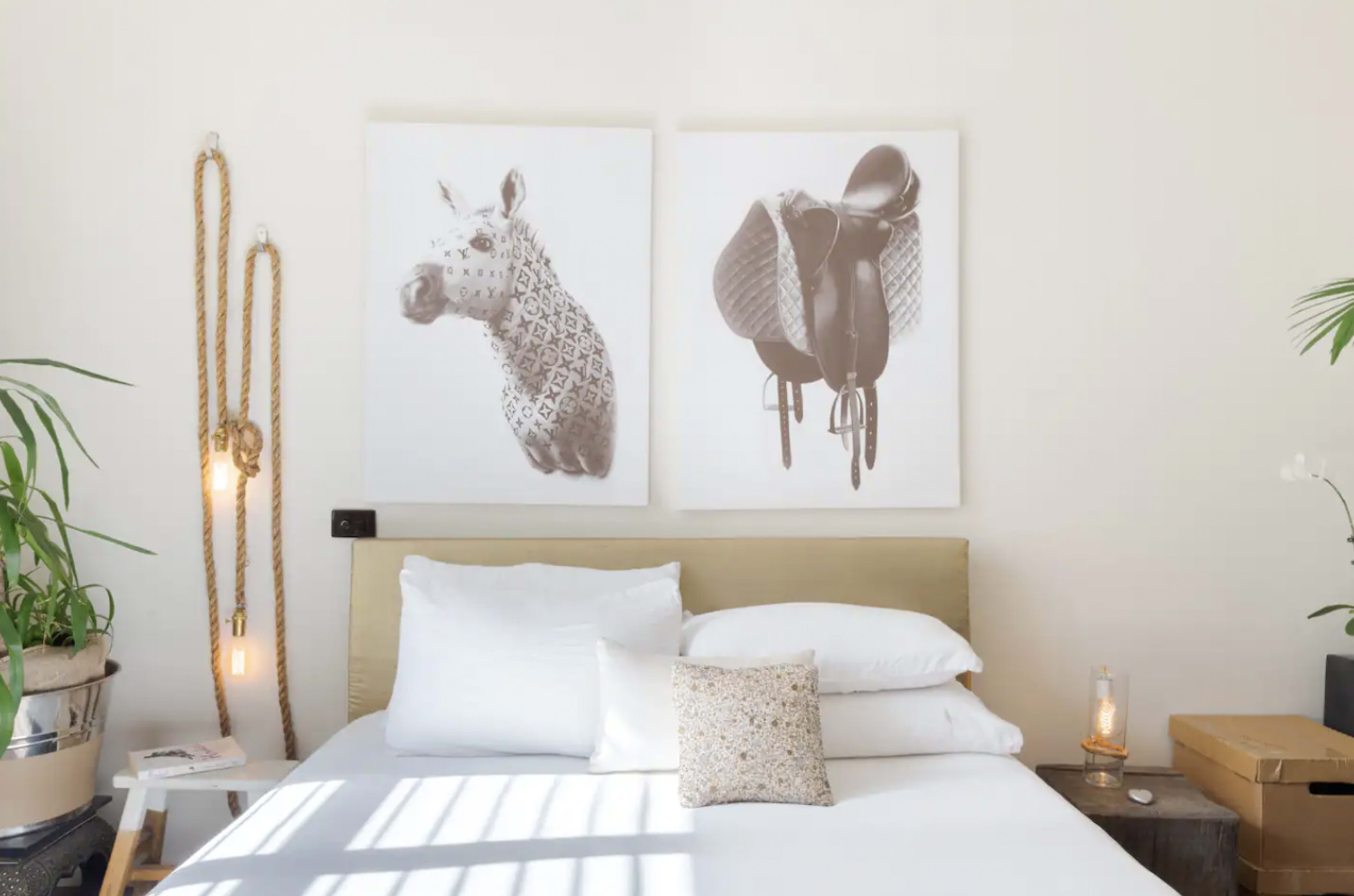 A modern bedroom with a bed adorned with white pillows, flanked by two framed artworks of a giraffe and a saddle on the wall.