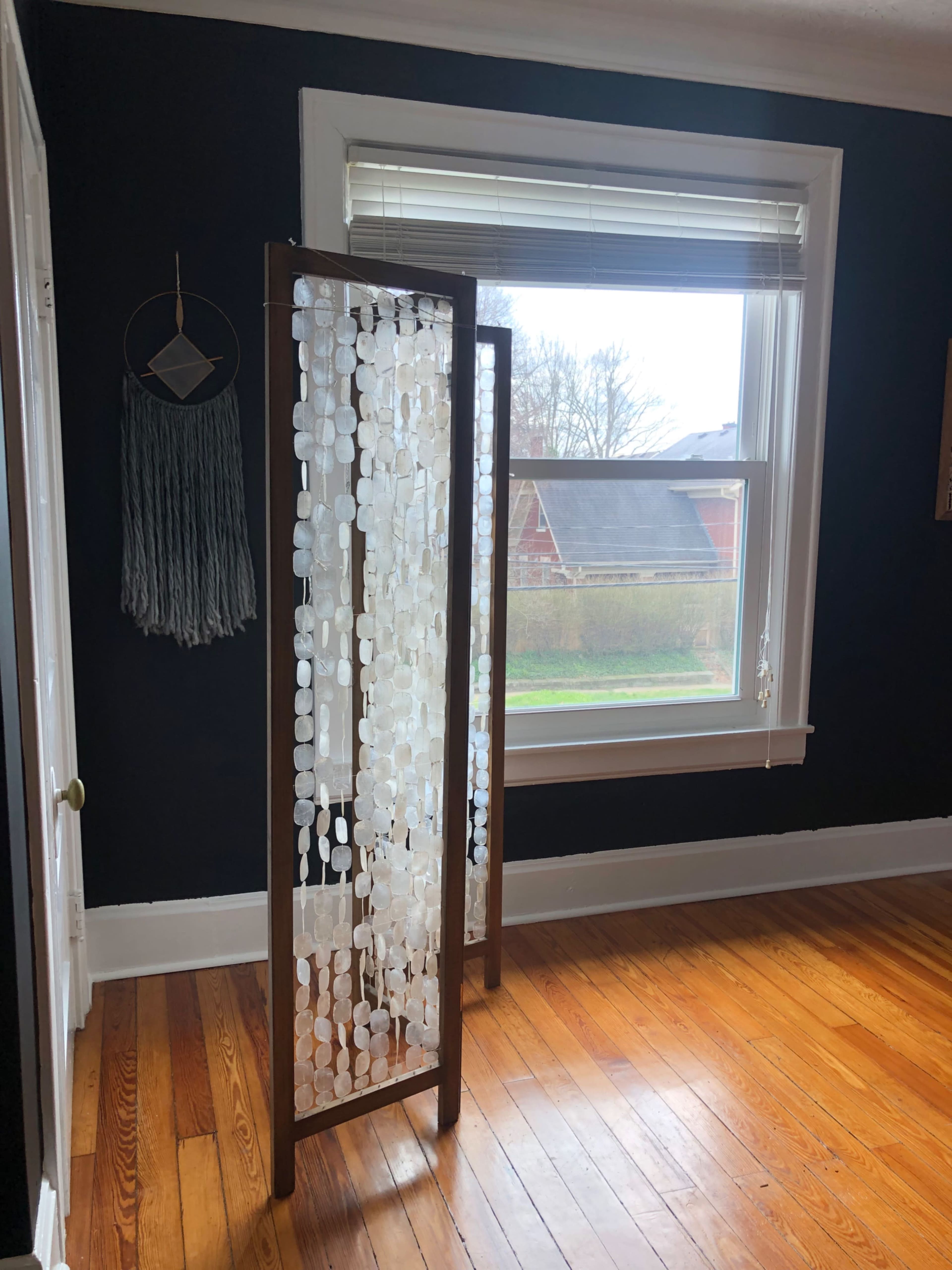 A room with dark walls features a wooden screen adorned with translucent discs, positioned near a window that overlooks a grassy area.
