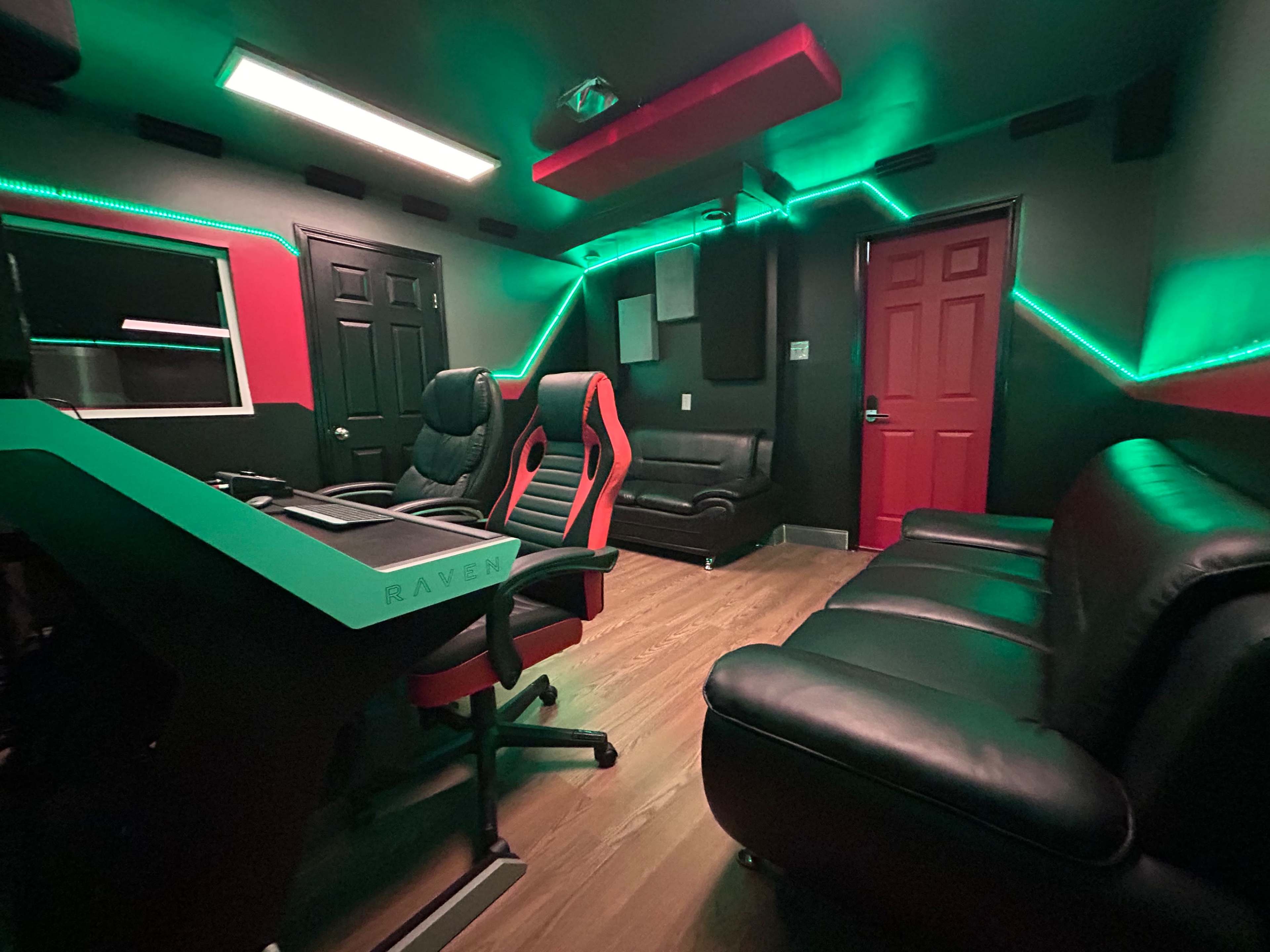 The image shows a modern recording studio with black walls, green and red LED lighting, a seating area, and a desk with computer equipment.