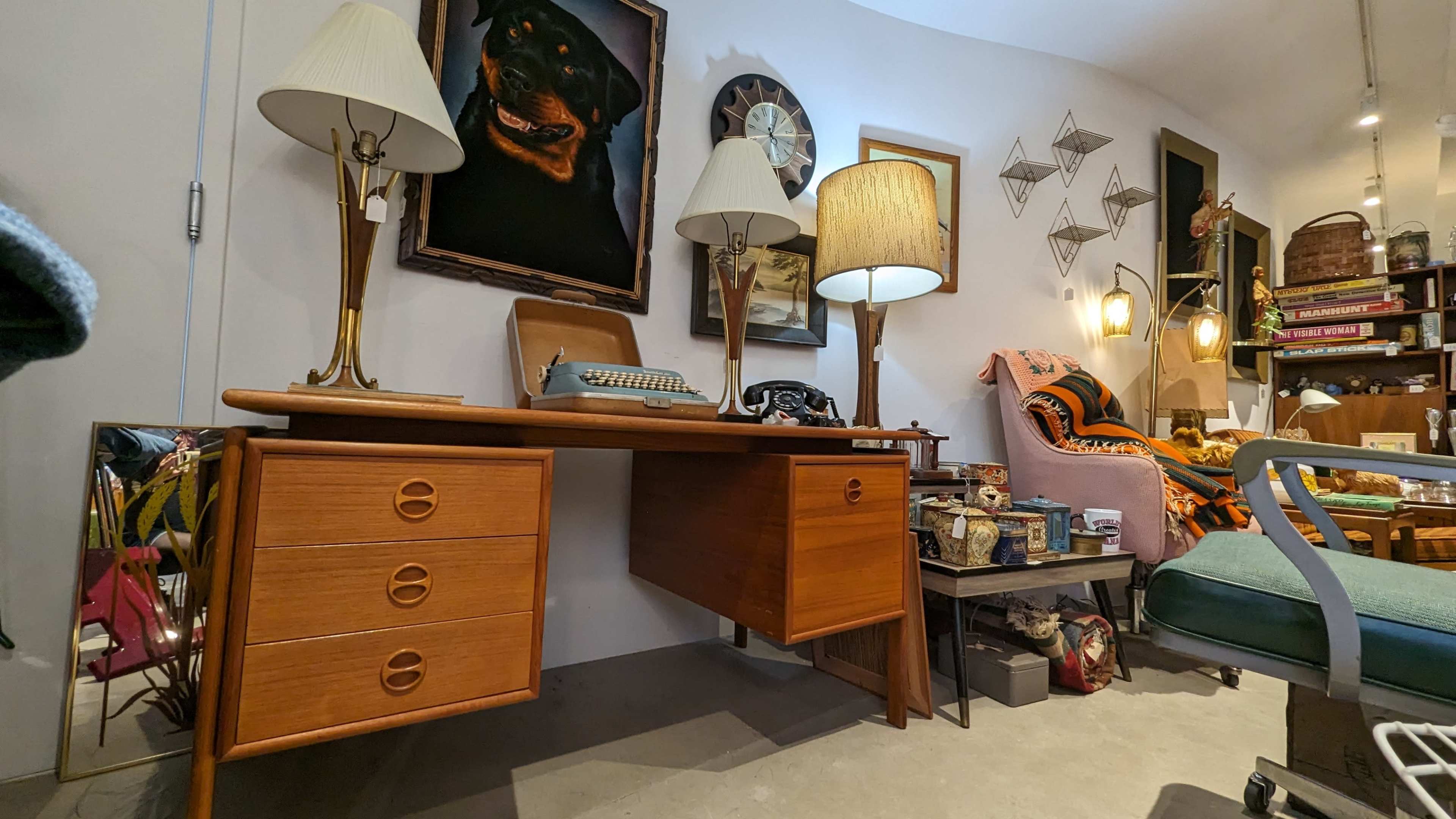A mid-century modern desk with drawers, a typewriter, and lamps, situated in a room decorated with various vintage items and art pieces.