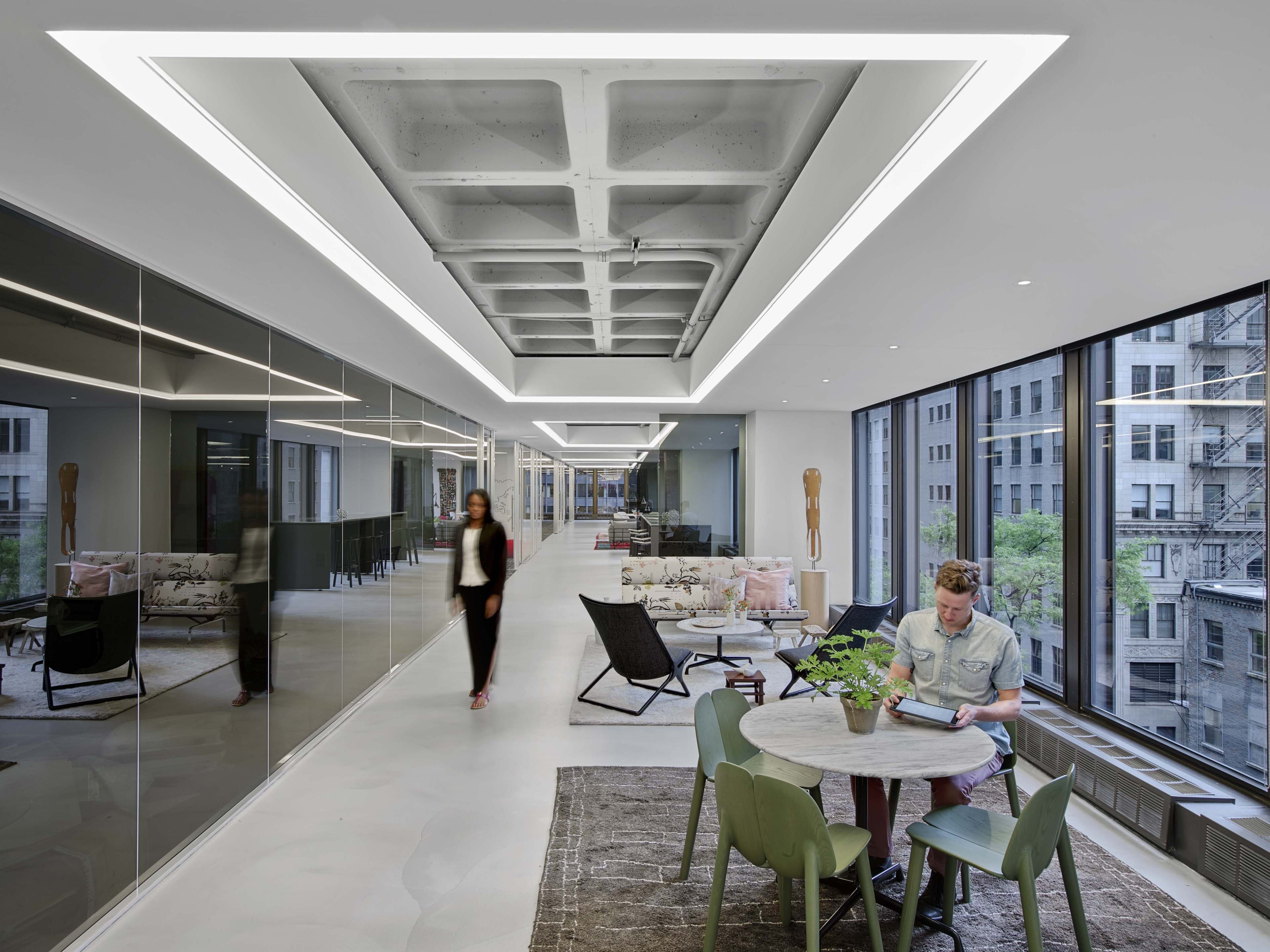 A modern office interior features a corridor with large windows, sleek furniture, and a person reading at a table while another walks by.