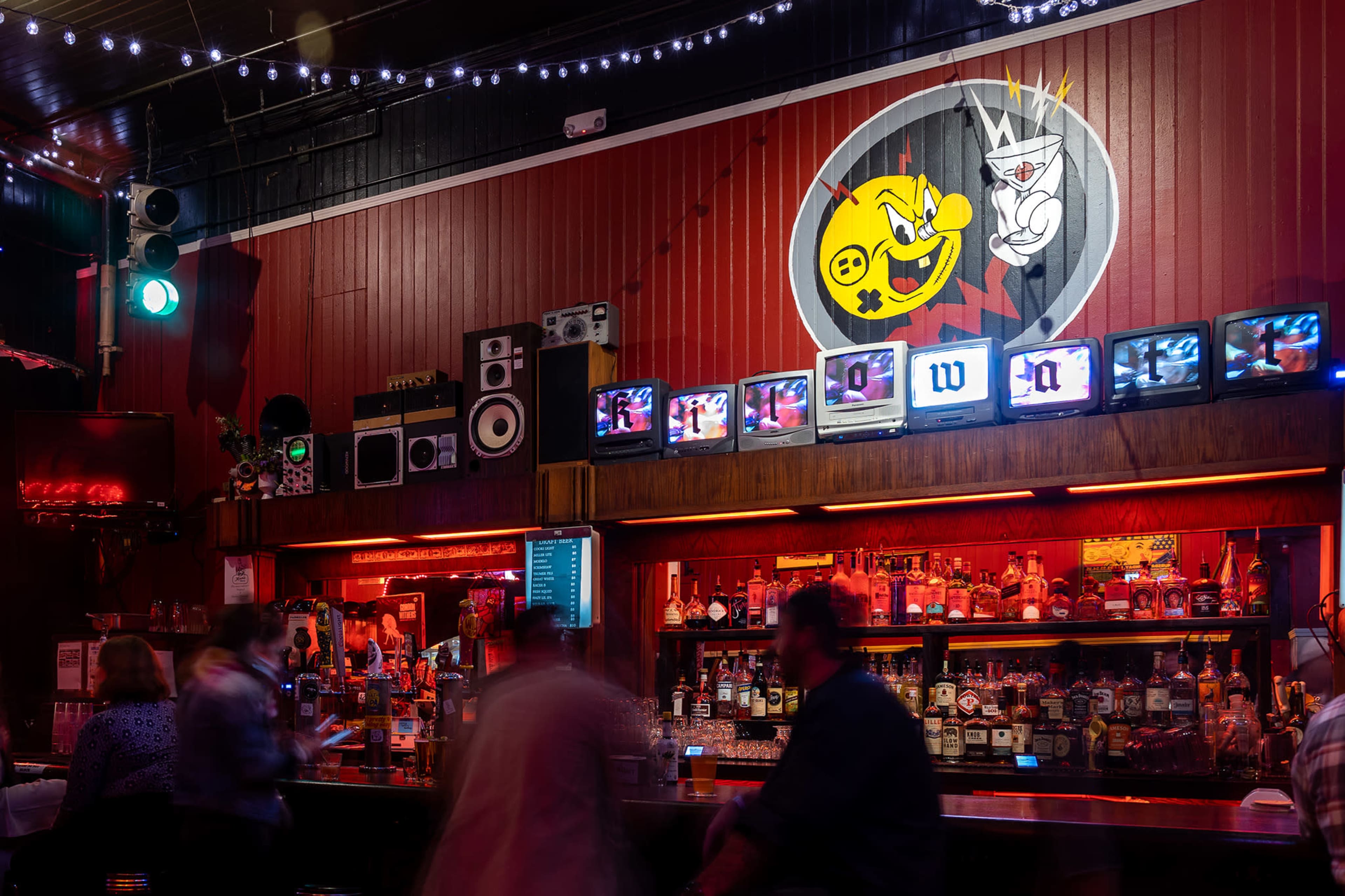 A dimly lit bar with a brightly painted mural of a mischievous character on the wall and multiple antique televisions displaying static along the bar counter.