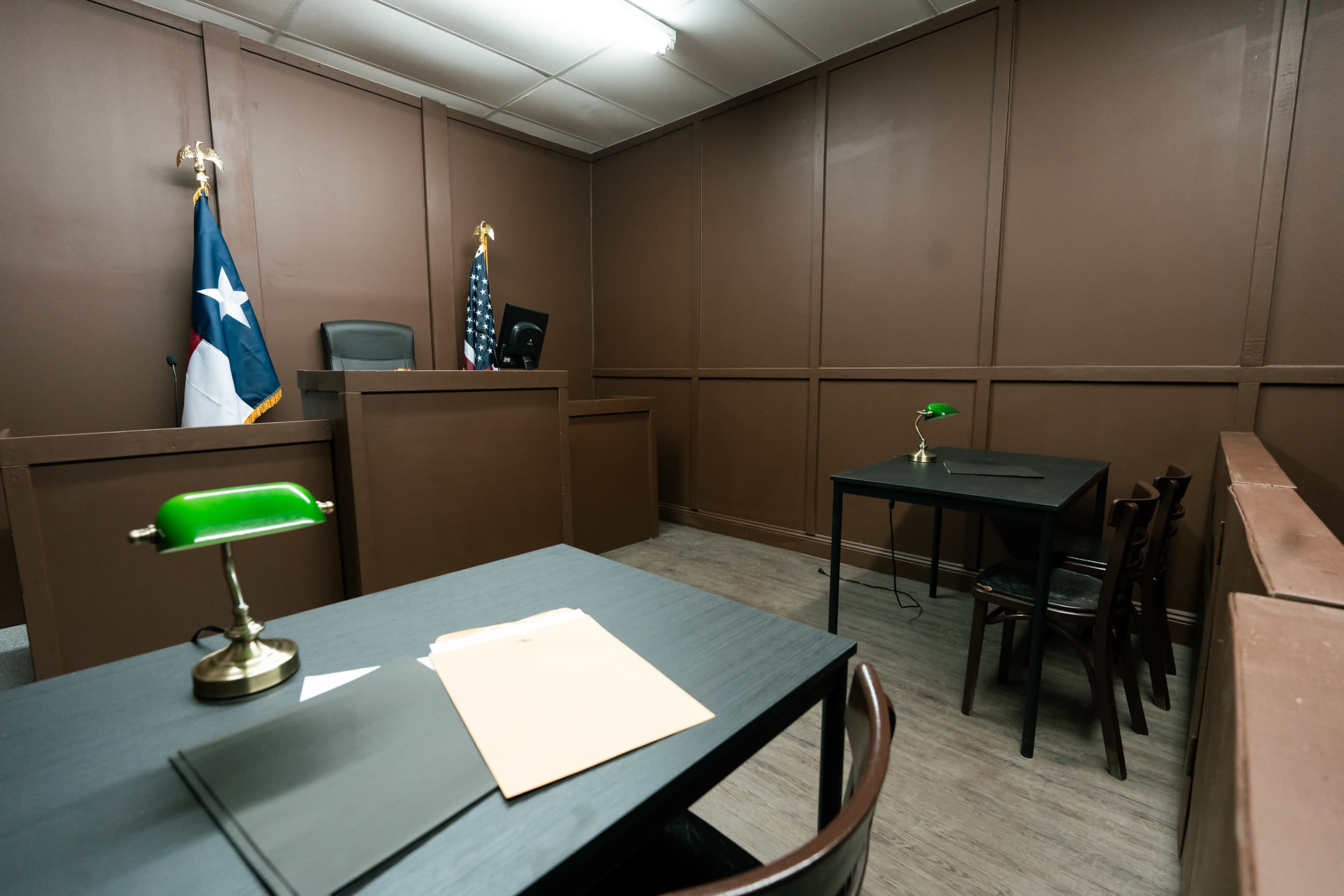 A small courtroom with a judge's bench, American and Texas flags, and two tables with chairs.
