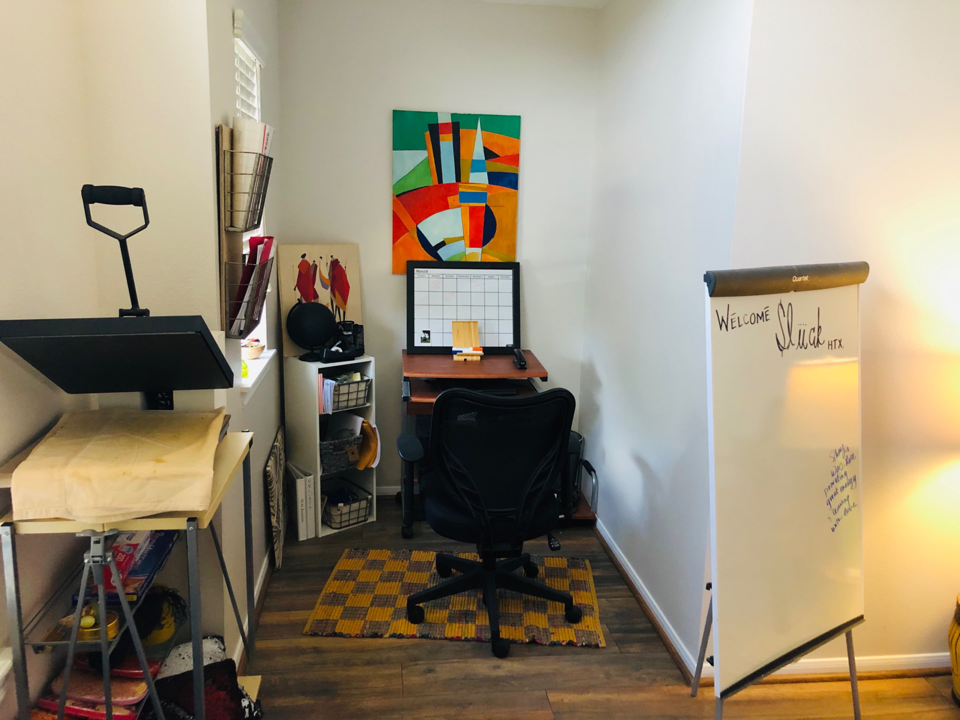 A small office space featuring a desk with a computer, an easel, a whiteboard, and colorful wall art.