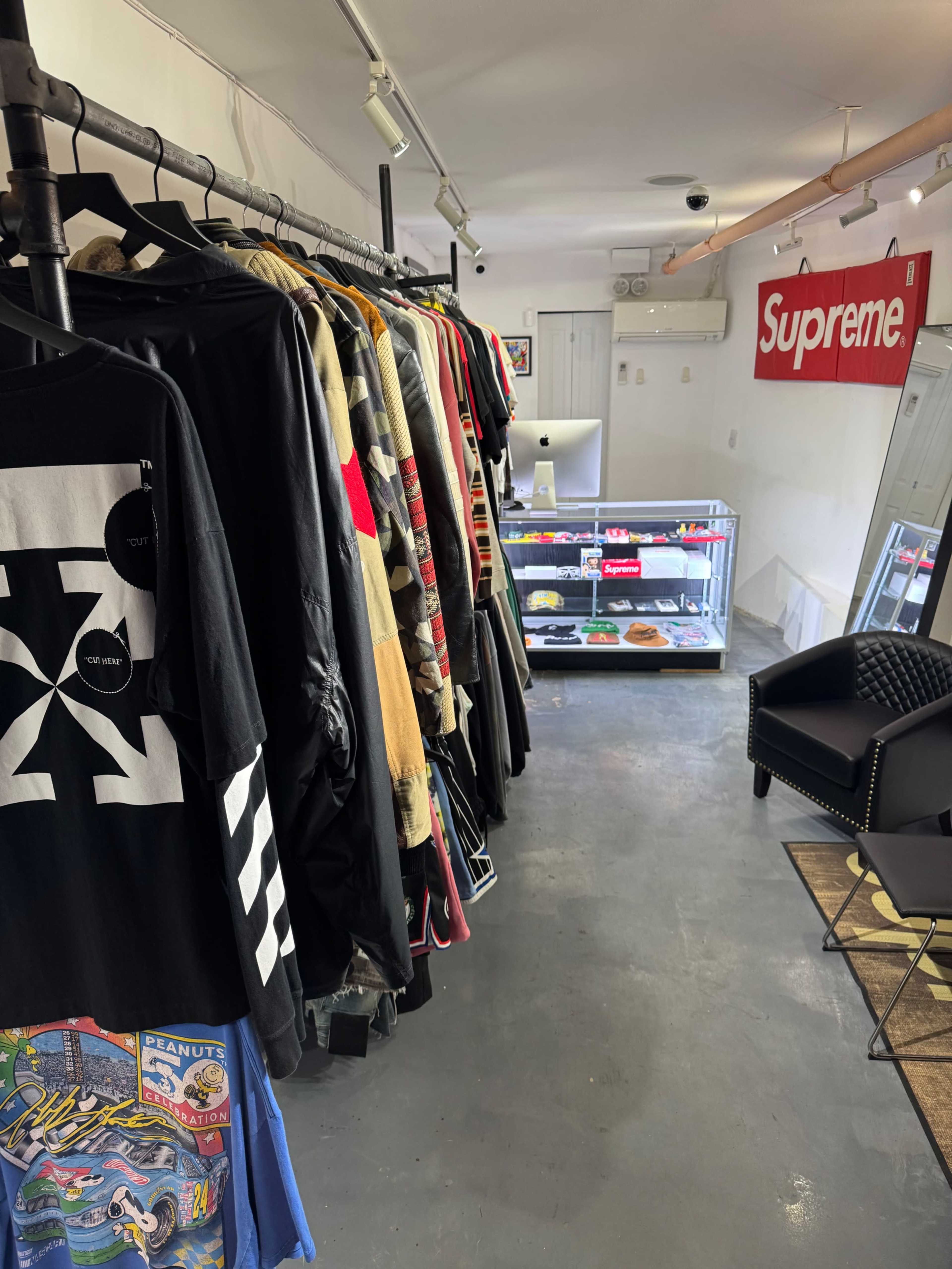 The image shows a retail space with a row of hanging clothing items on racks, a display case filled with merchandise, and a chair in the corner.