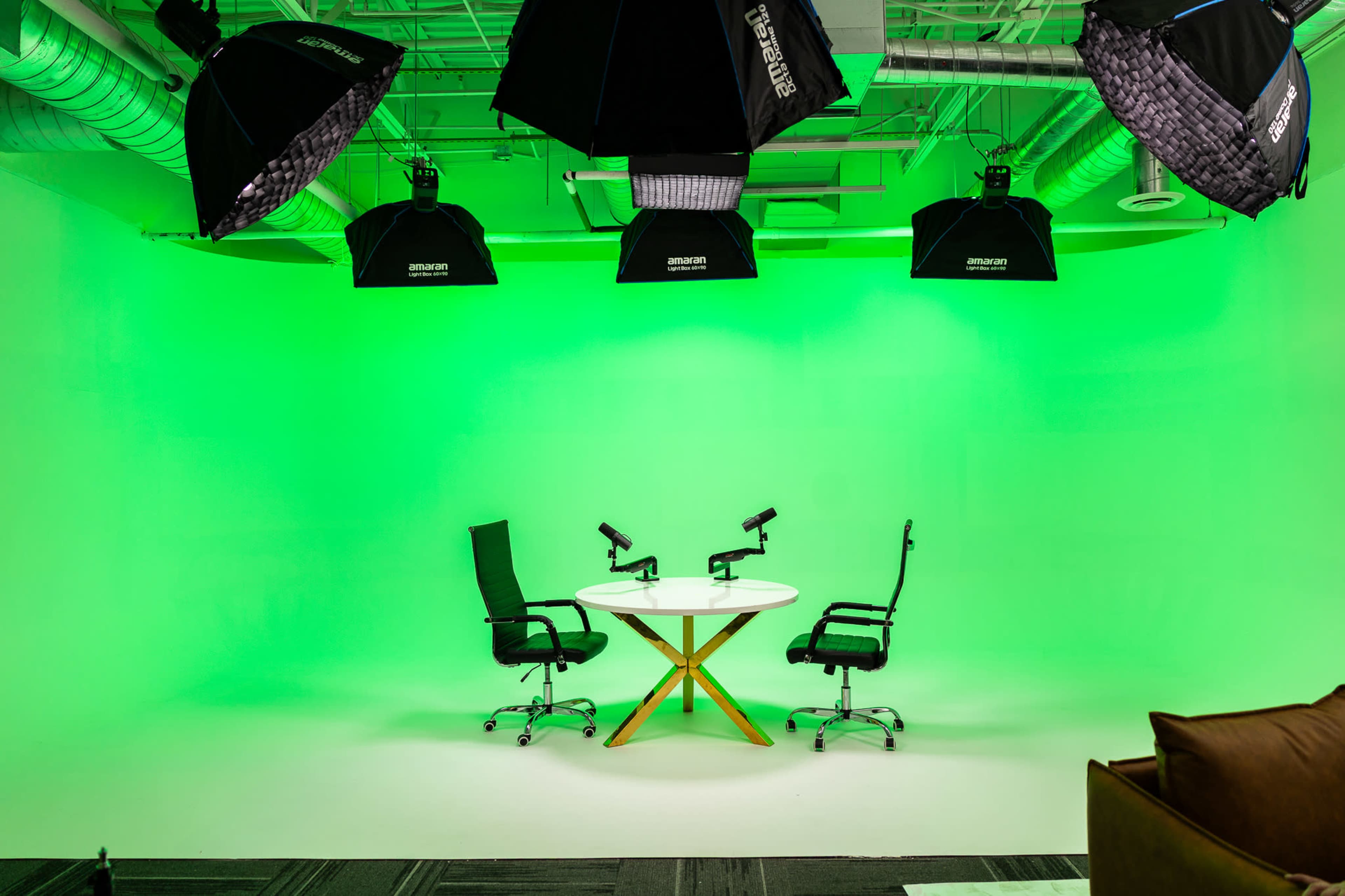 The image shows a green-screen studio featuring a round table with two chairs positioned opposite each other under several overhead lights.