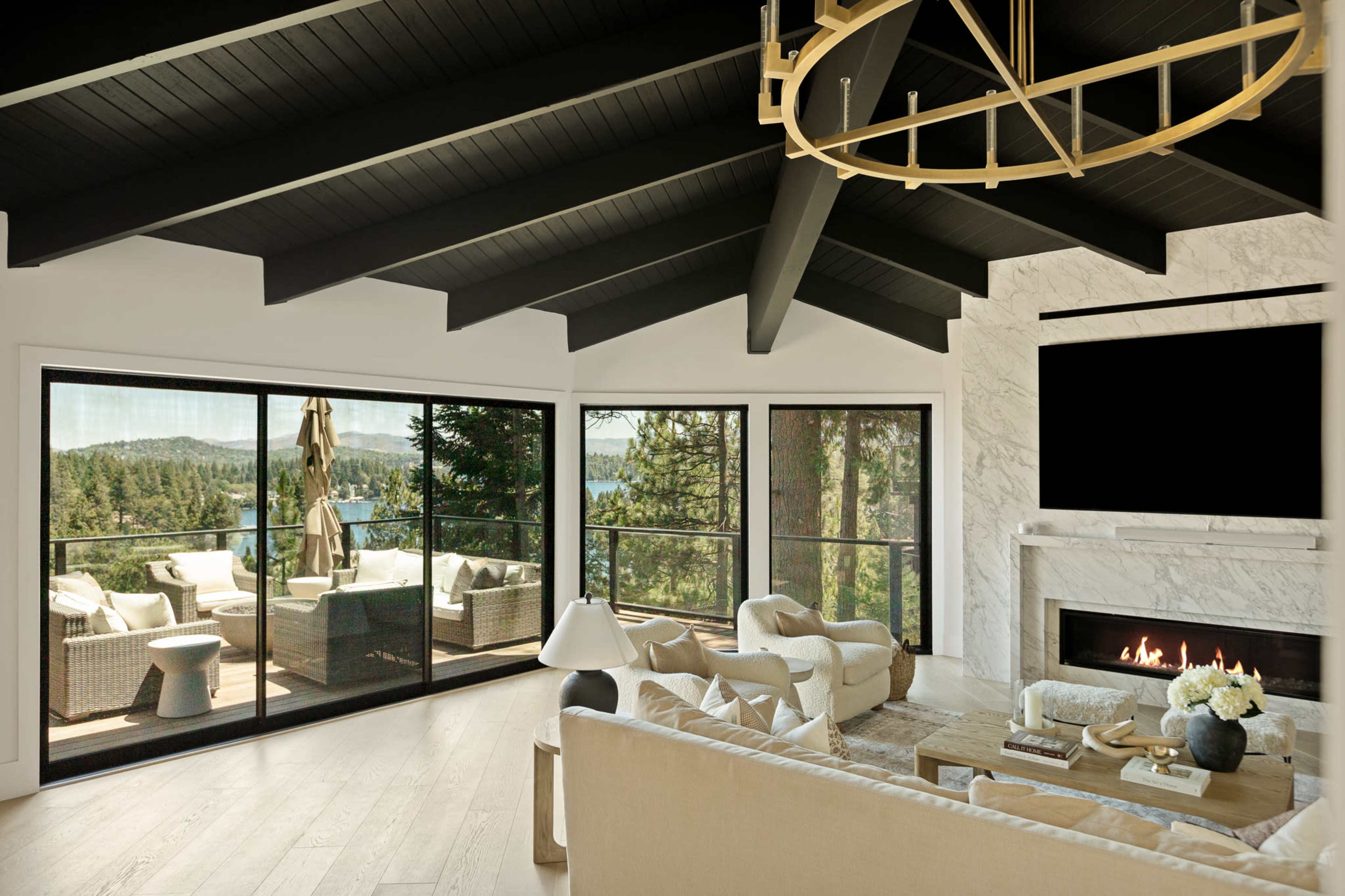 The image shows a modern living room with high vaulted ceilings, large windows overlooking a wooded landscape, and a minimalist fireplace.
