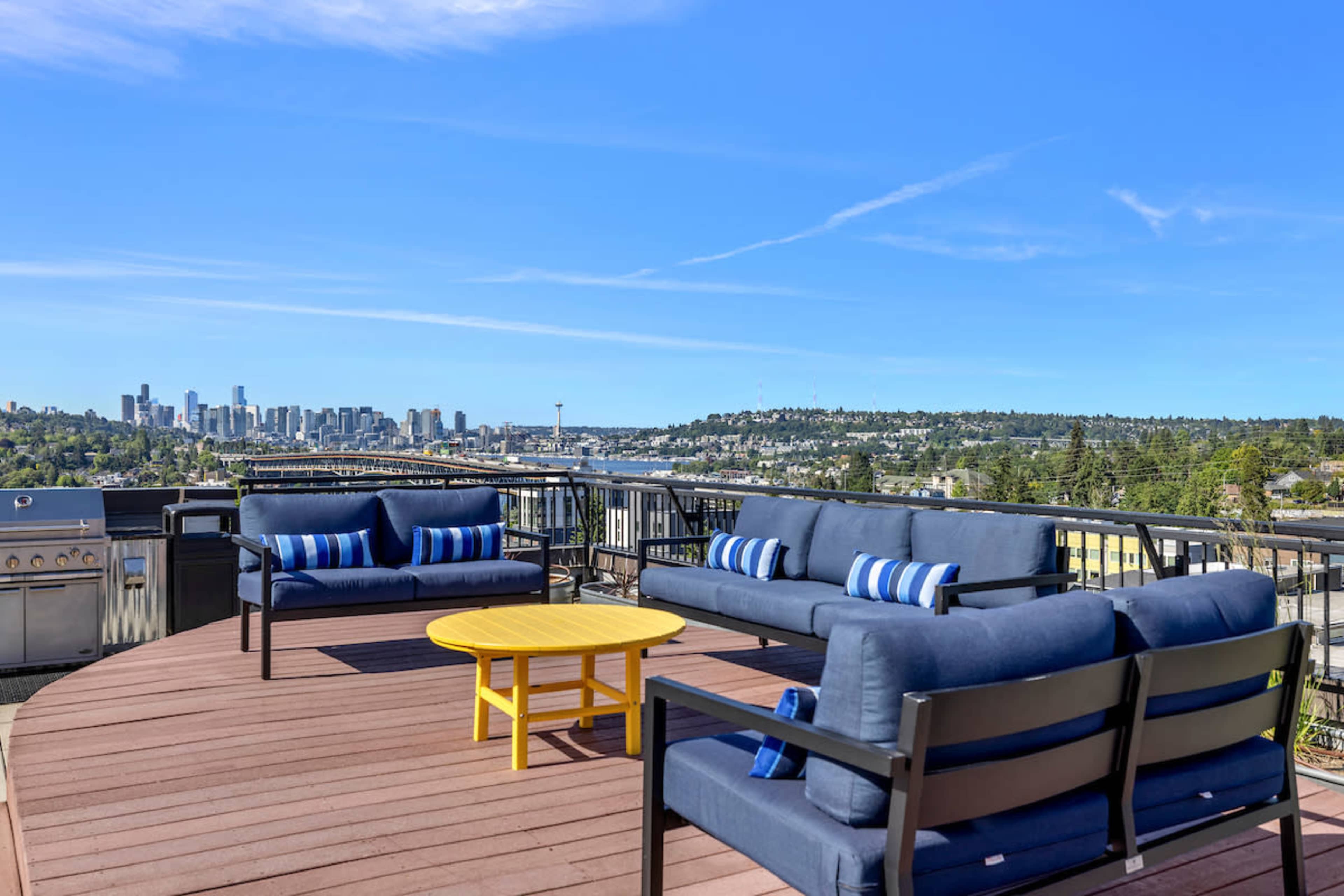 The image shows a rooftop terrace with outdoor furniture, including blue sofas and a yellow coffee table, overlooking a city skyline on a clear day.