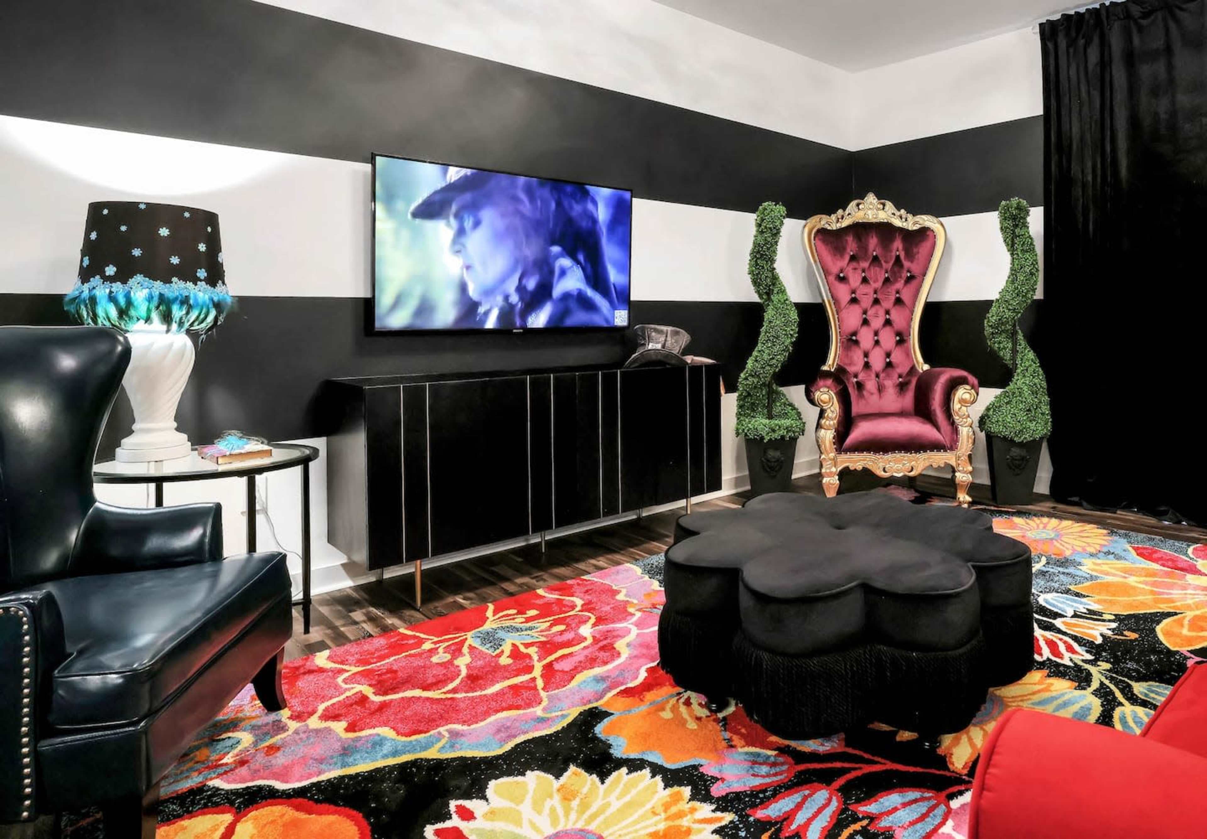 A living room featuring a black and white striped wall, a large television mounted on the wall, a purple velvet armchair, a black round coffee table, and a colorful floral area rug.