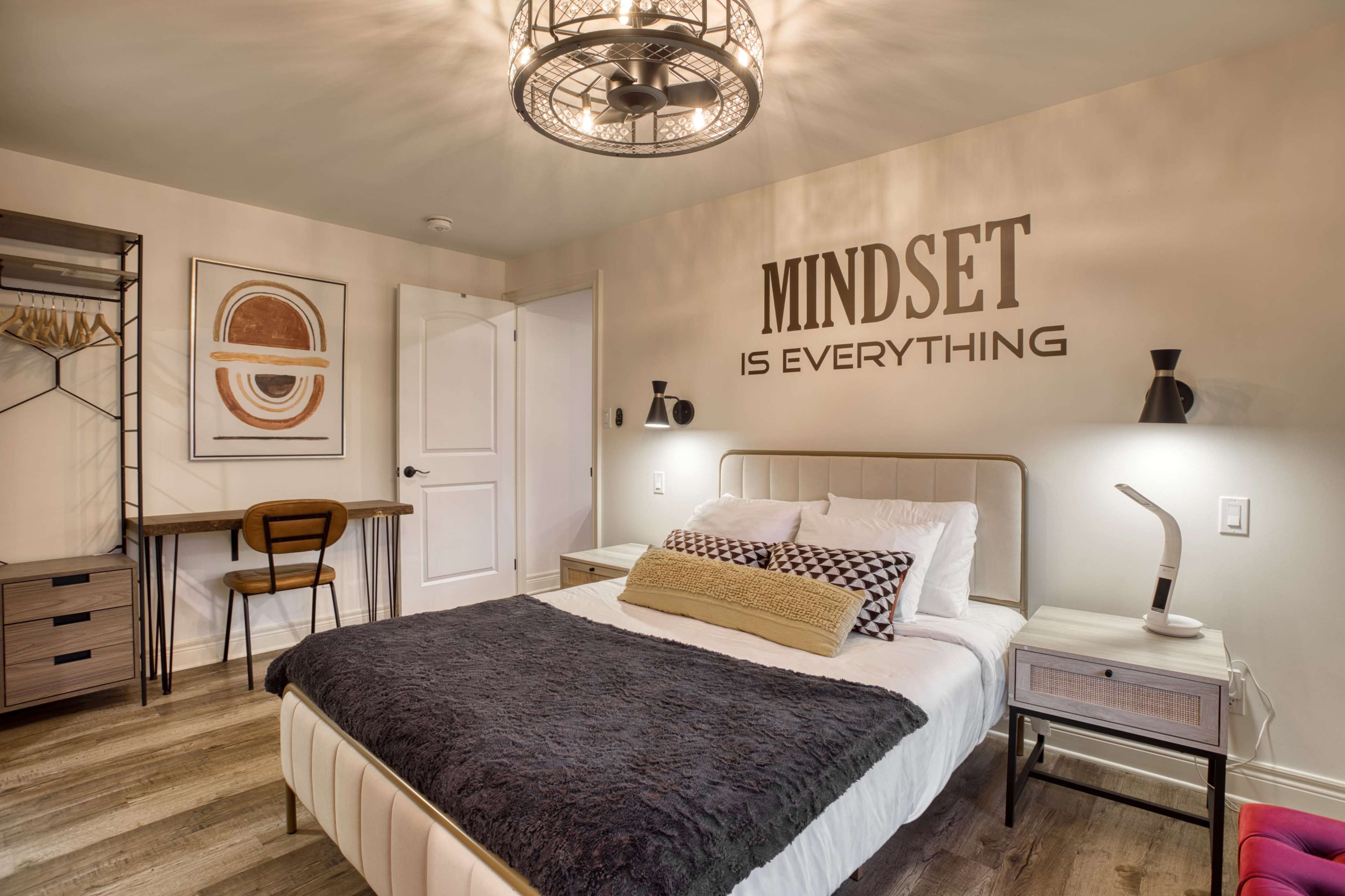 The image shows a modern bedroom featuring a bed with a gray throw, a nightstand with a lamp, a wall art piece, and a desk with a chair.