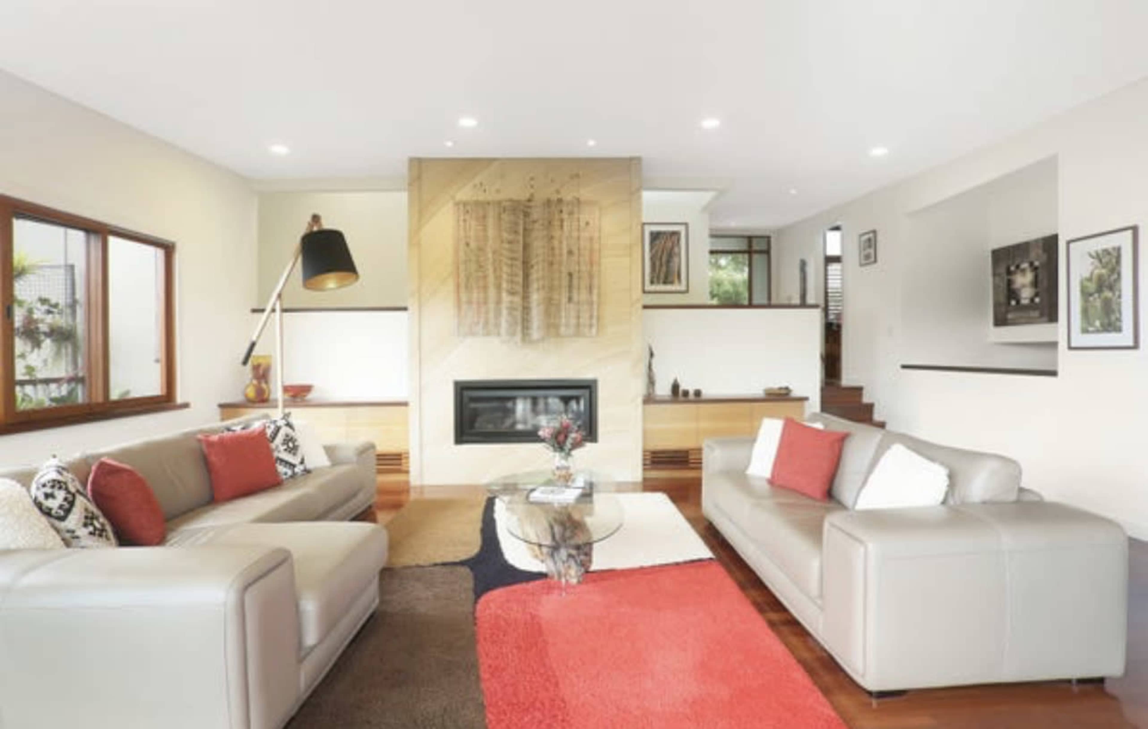 A modern living room with two sofas facing a central glass coffee table, a fireplace in the background, and a combination of wood and carpet flooring.