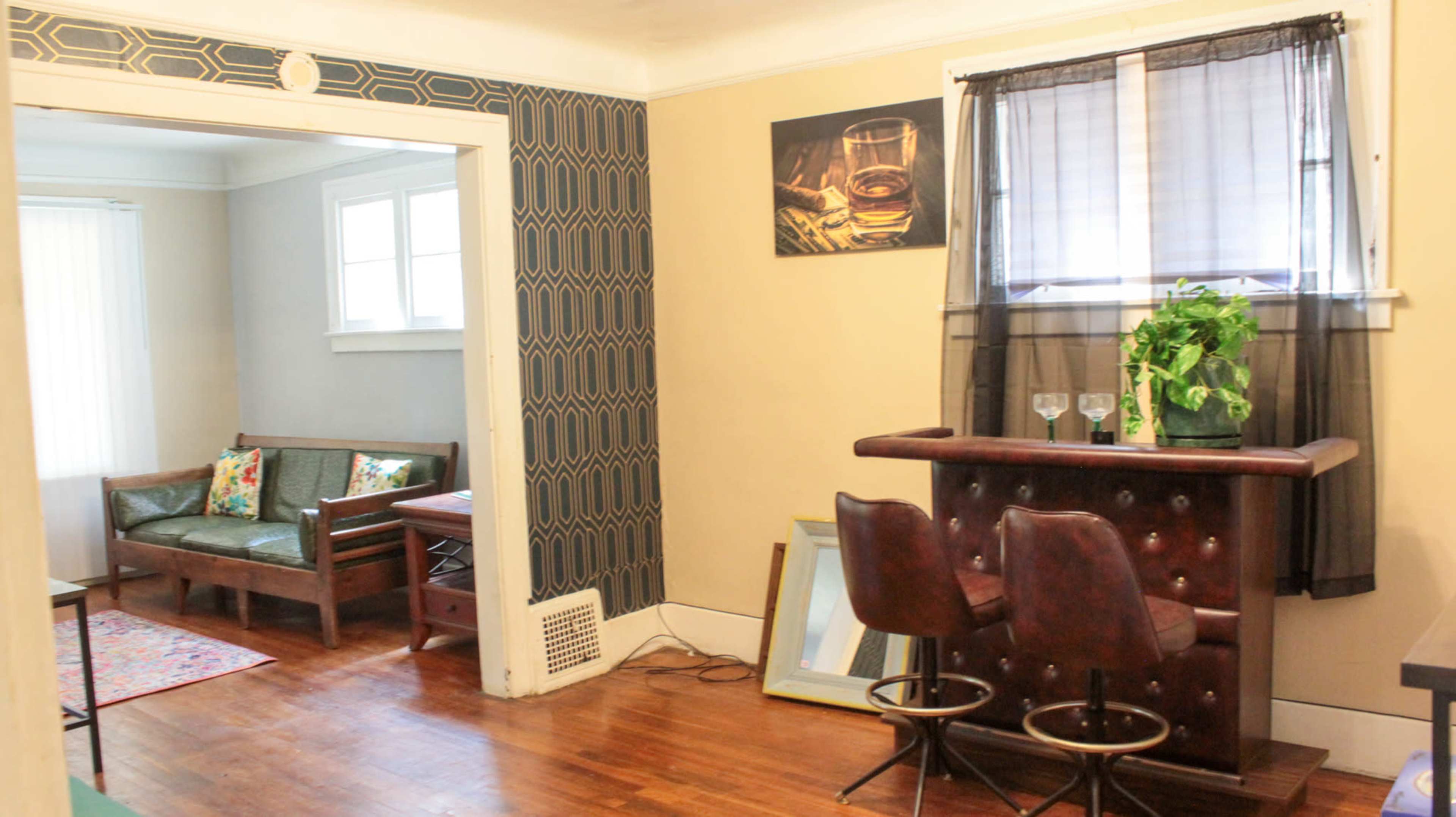 A room with a small bar area featuring two stools, a potted plant, and a view into a separate living space with a couch and decorative pillows.