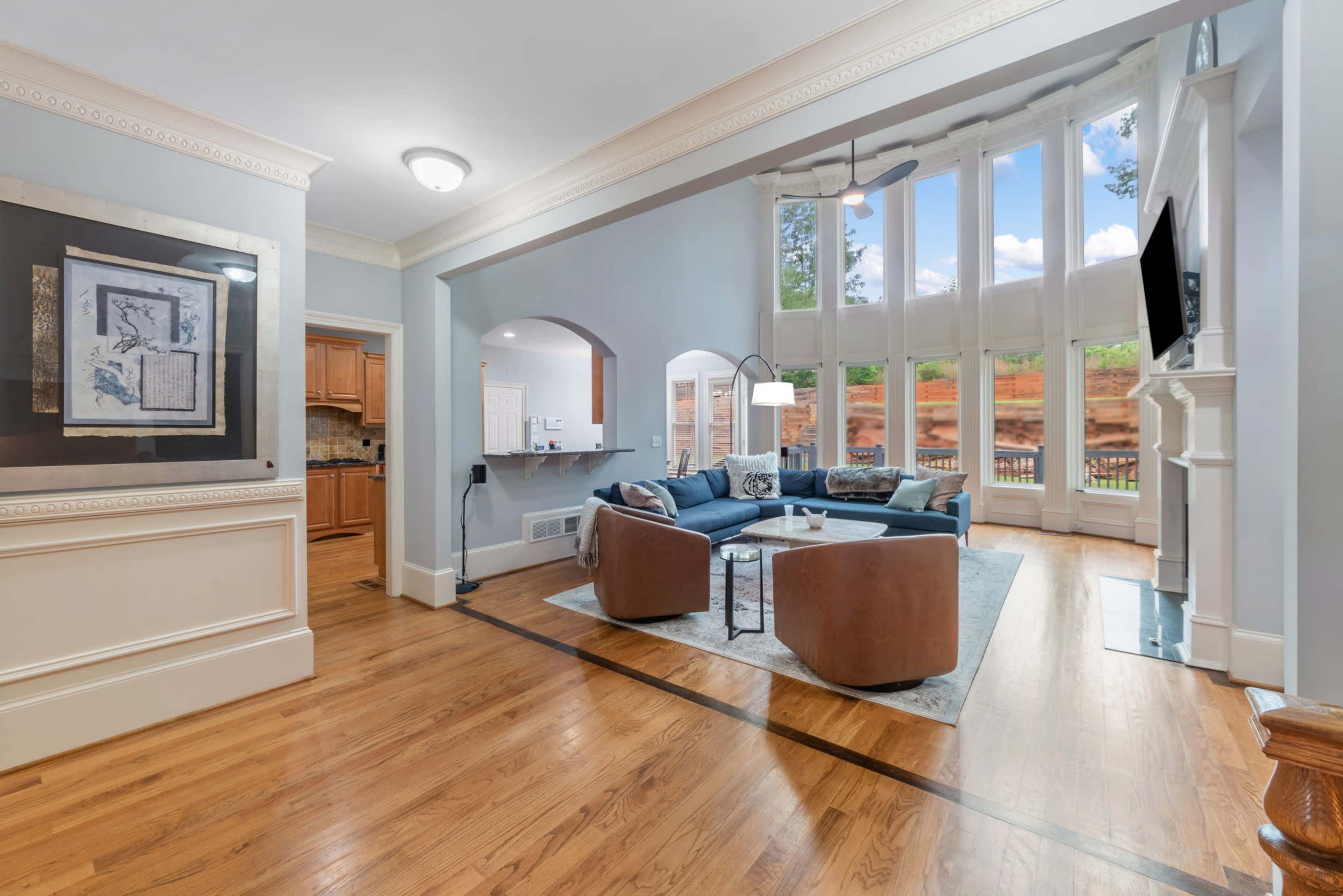 A spacious living room features large windows, a blue sectional sofa, and hardwood flooring, with a fireplace and a kitchen visible in the background.