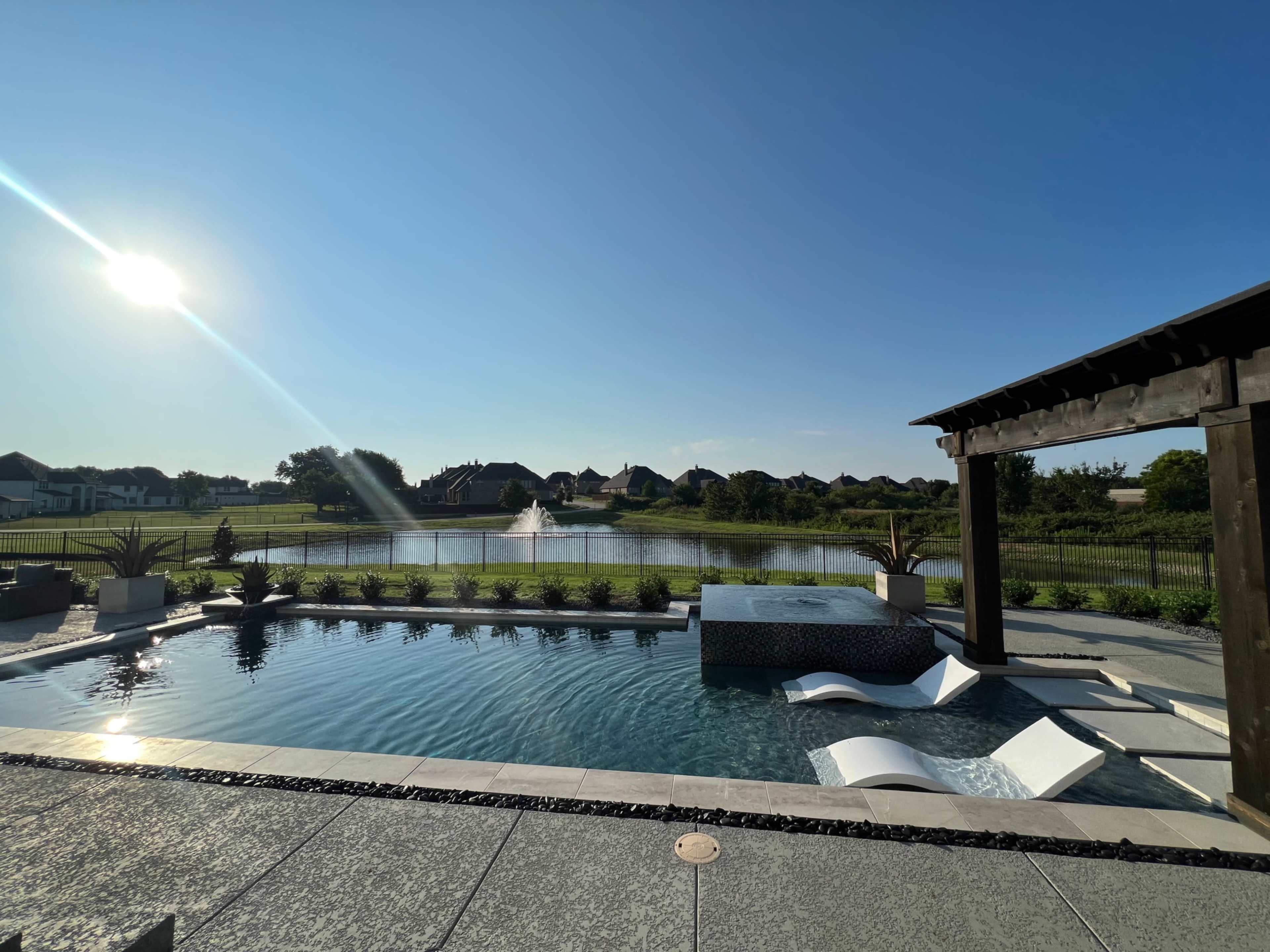 The image shows a modern backyard with a swimming pool, a spa area, and a scenic view of a pond and homes in the background under a clear blue sky.